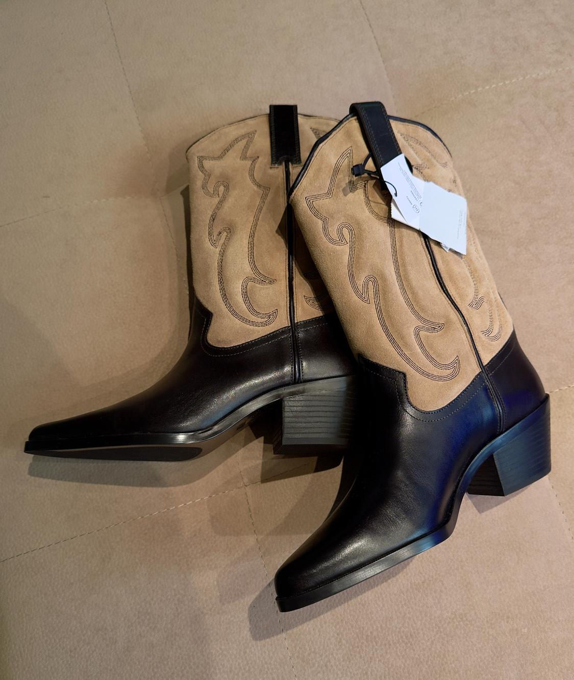 Pair of cowboy-style boots against a beige background. They have black soles, beige suede shafts with embroidery, chunky heels and a white label.