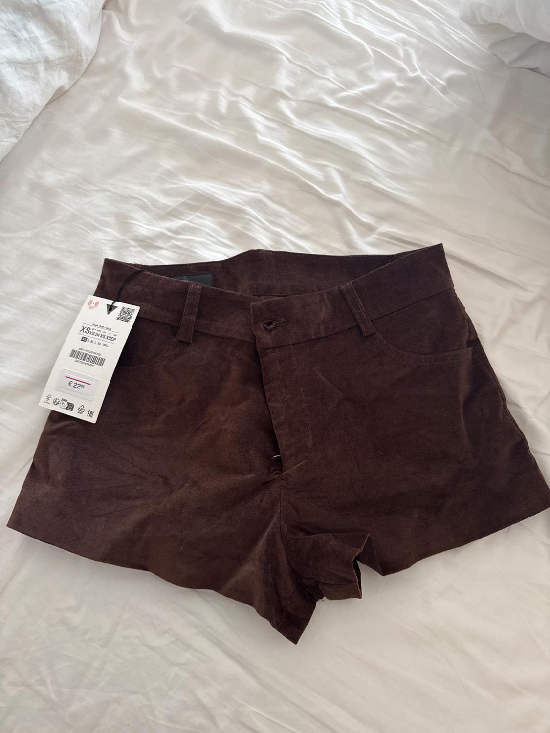 Brown velvet shorts on a white bed, with a tag showing size XS and price €22.