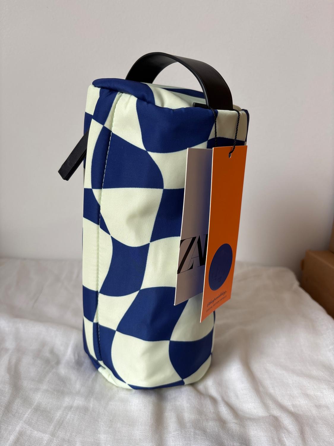 Cylindrical bag with a blue and white checkered print, black handle, and an orange tag with the text "ZA". White background.