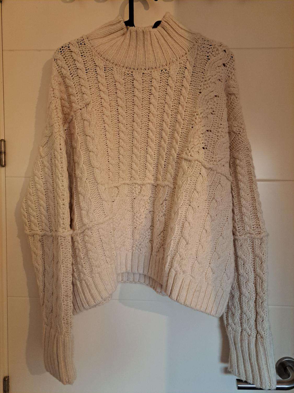 Chunky cream cable-knit jumper hanging on a hanger over a white door, with a high neck.
