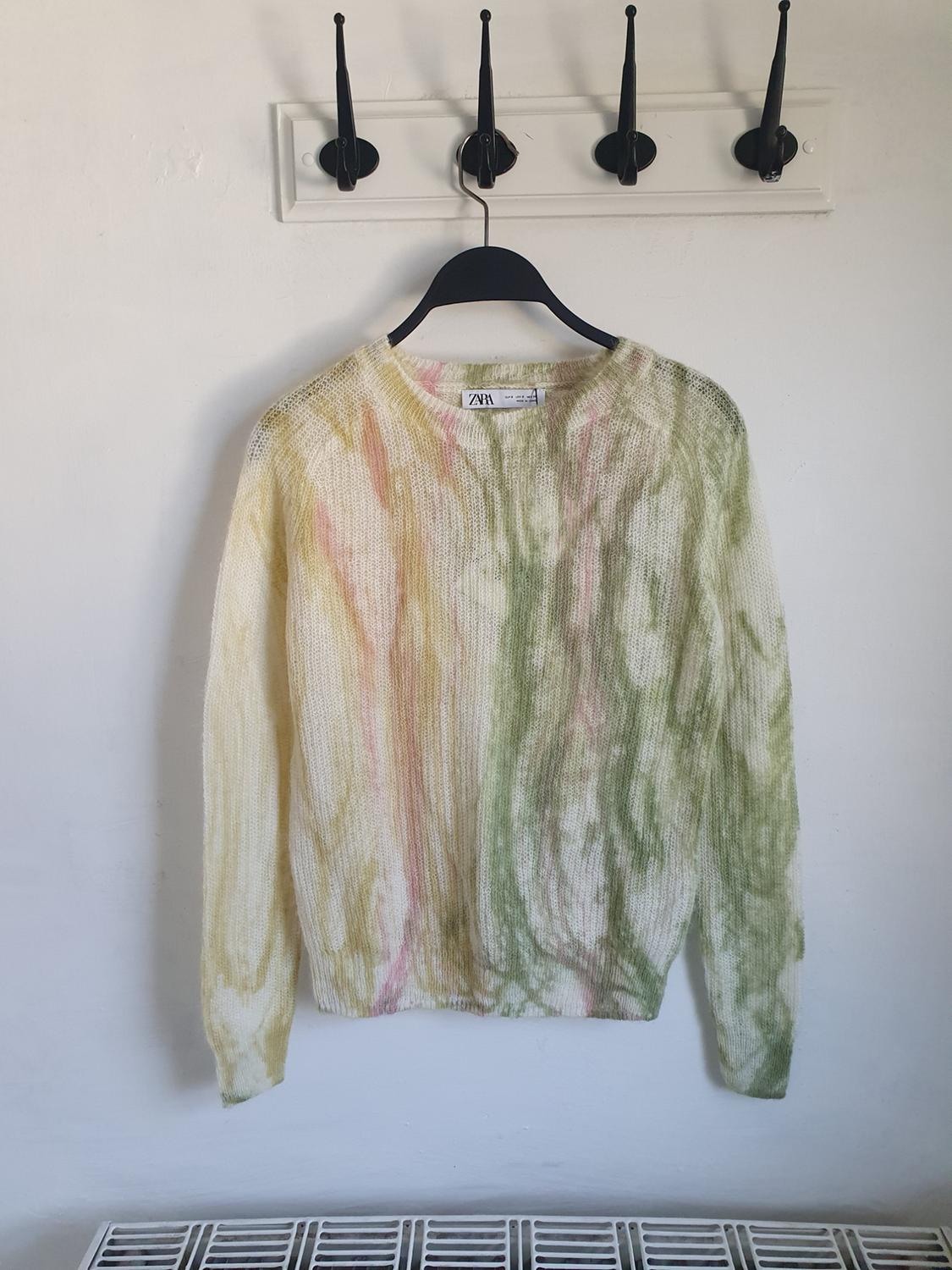 Knitted jumper with a green and pink patterned print, hanging on a black hanger against a white background with hooks.