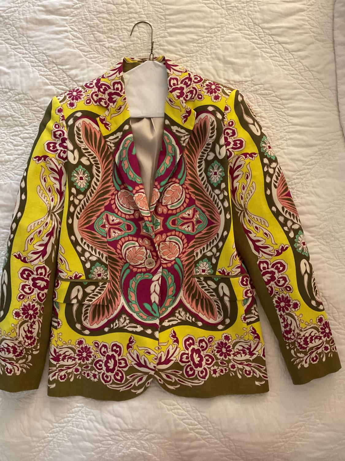 Blazer with a striking ornamental print in yellow, fuchsia, and green, hanging on a hanger over a white quilted bedspread.