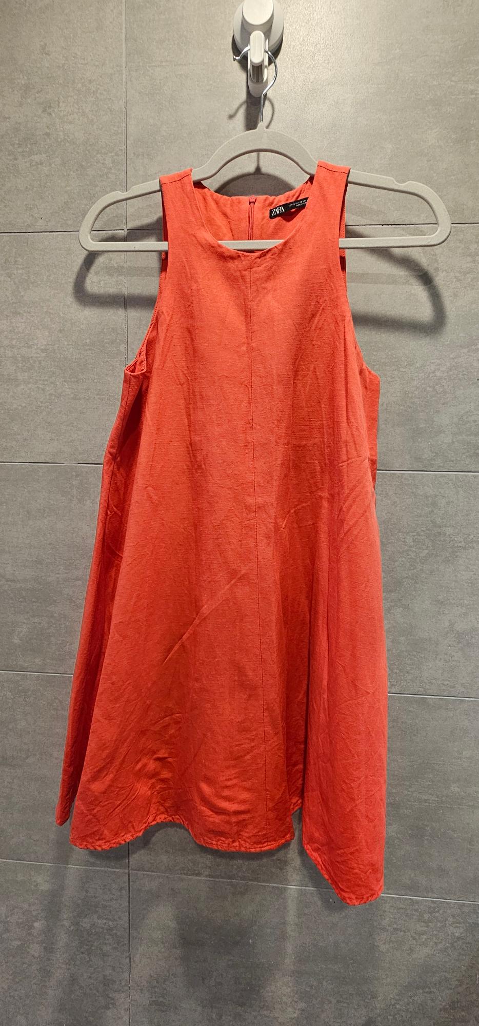 Sleeveless red dress hanging on a grey hanger against a grey tiled wall. Label visible on the collar.