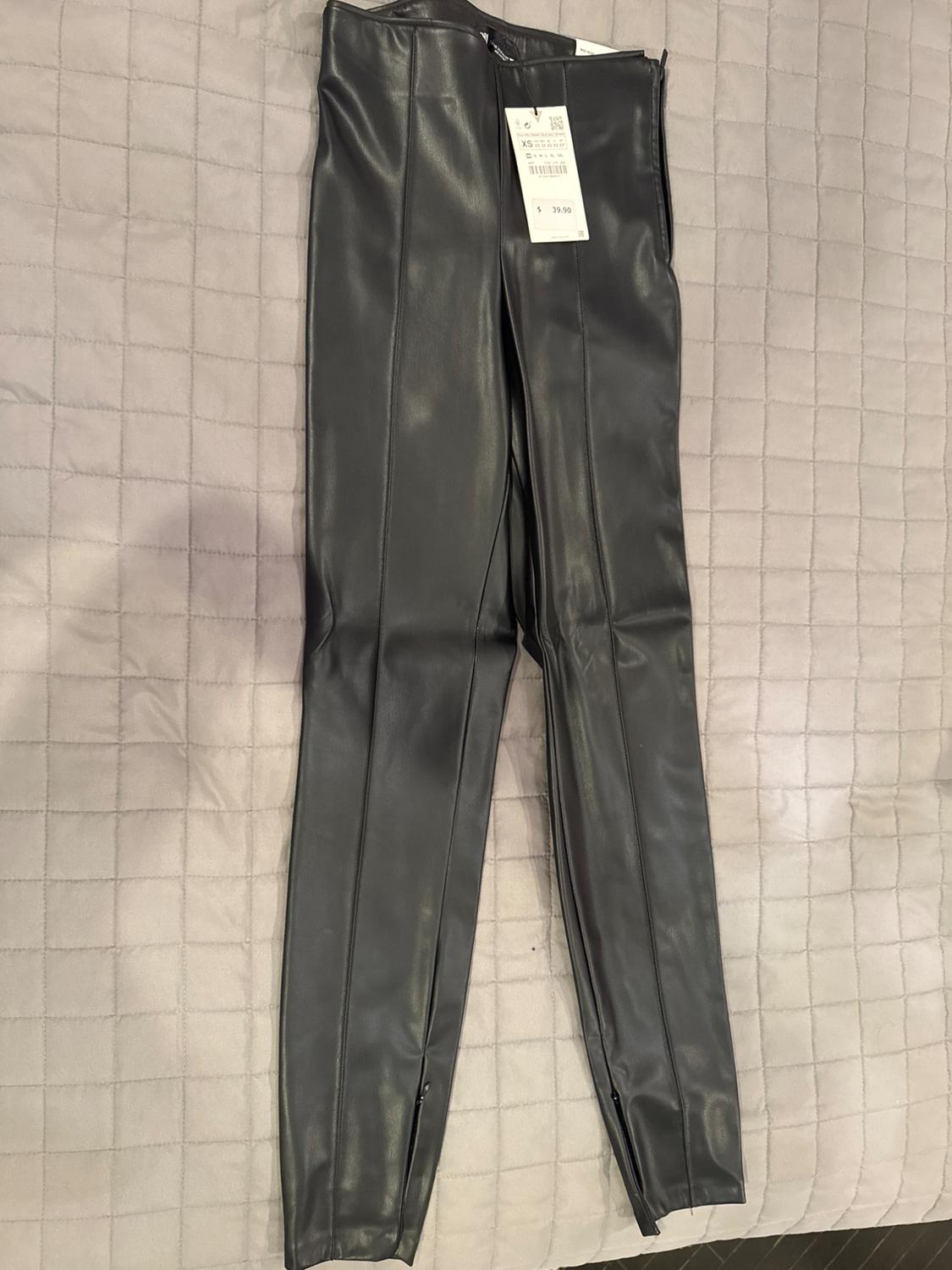 Front view of black pants with visible tag, vertical seams, and ankle zippers.