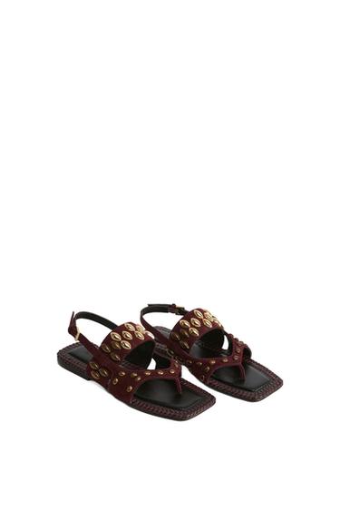 Burgundy leather sandals with gold studs and a black sole, on a white background. Elegant and modern design.