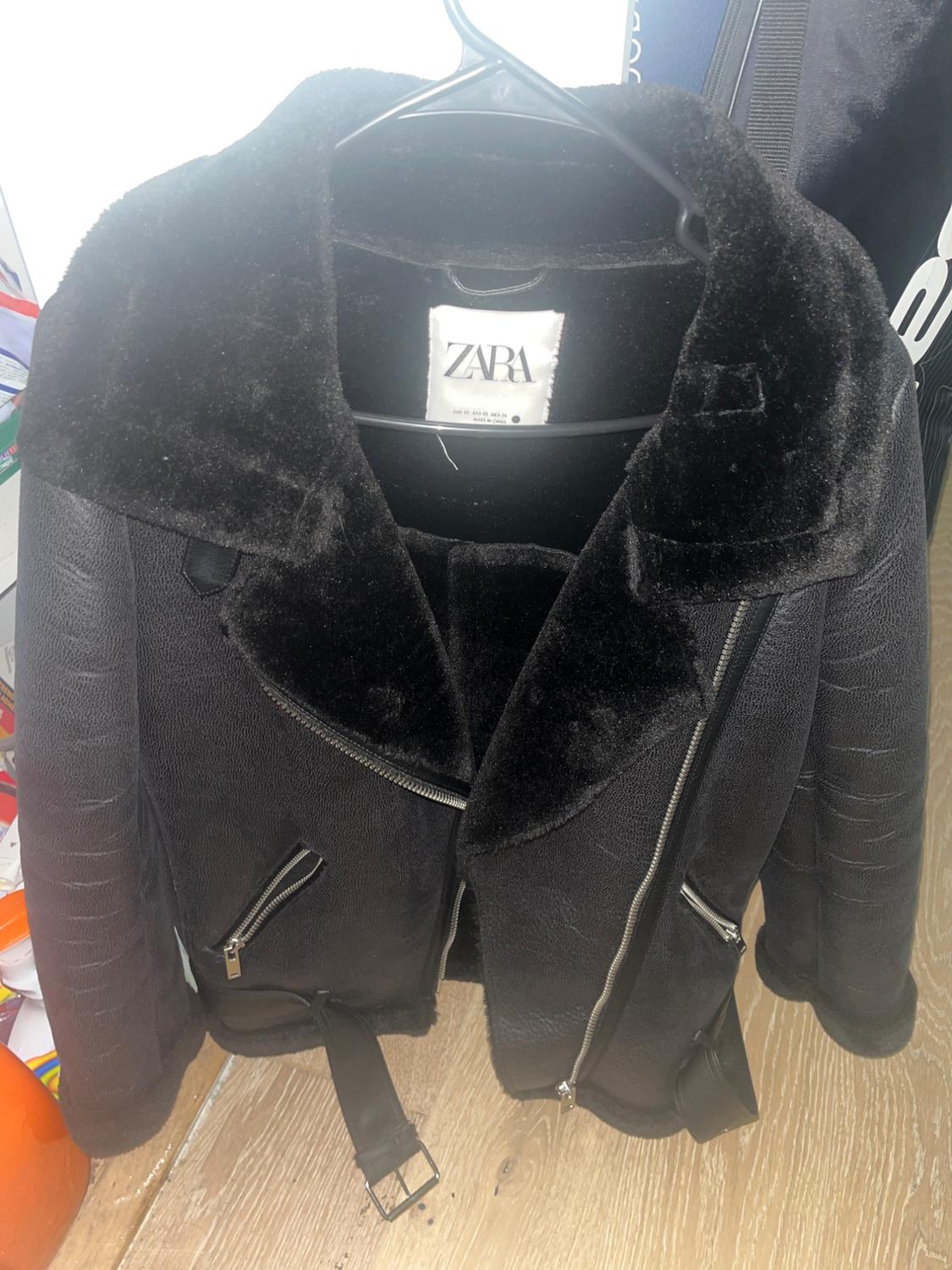 Black faux leather jacket with a plush collar and zippers, hanging on a hanger. Visible tag: ZARA.