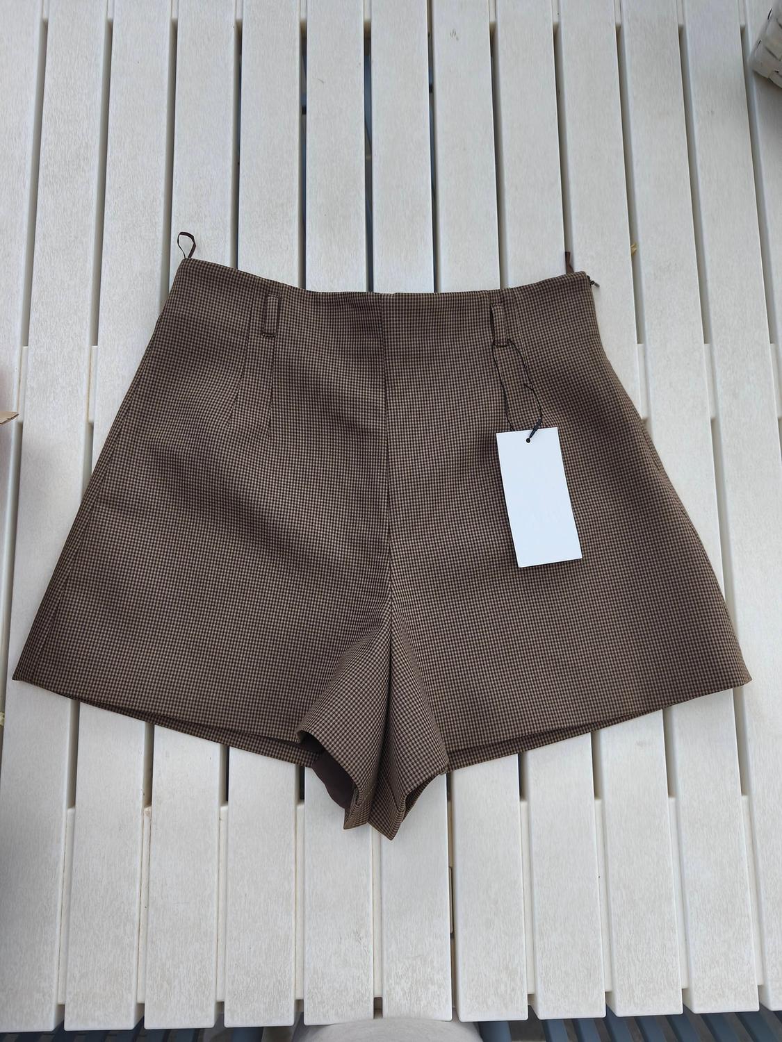 Brown shorts with a small check print, hung over a white wooden surface, with a white tag attached.