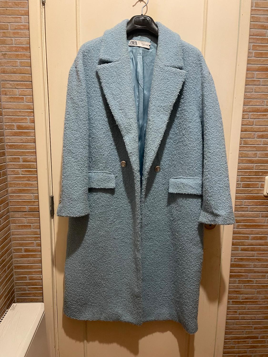 Light blue wool coat hanging on a door, with wide lapels and two buttons. Visible label with the ZARA brand. Tiled background.