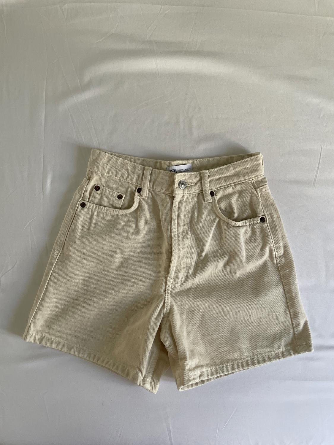 Light beige high-waisted denim shorts with five pockets, laid out on a white fabric. Inner label bearing the text ZARA.