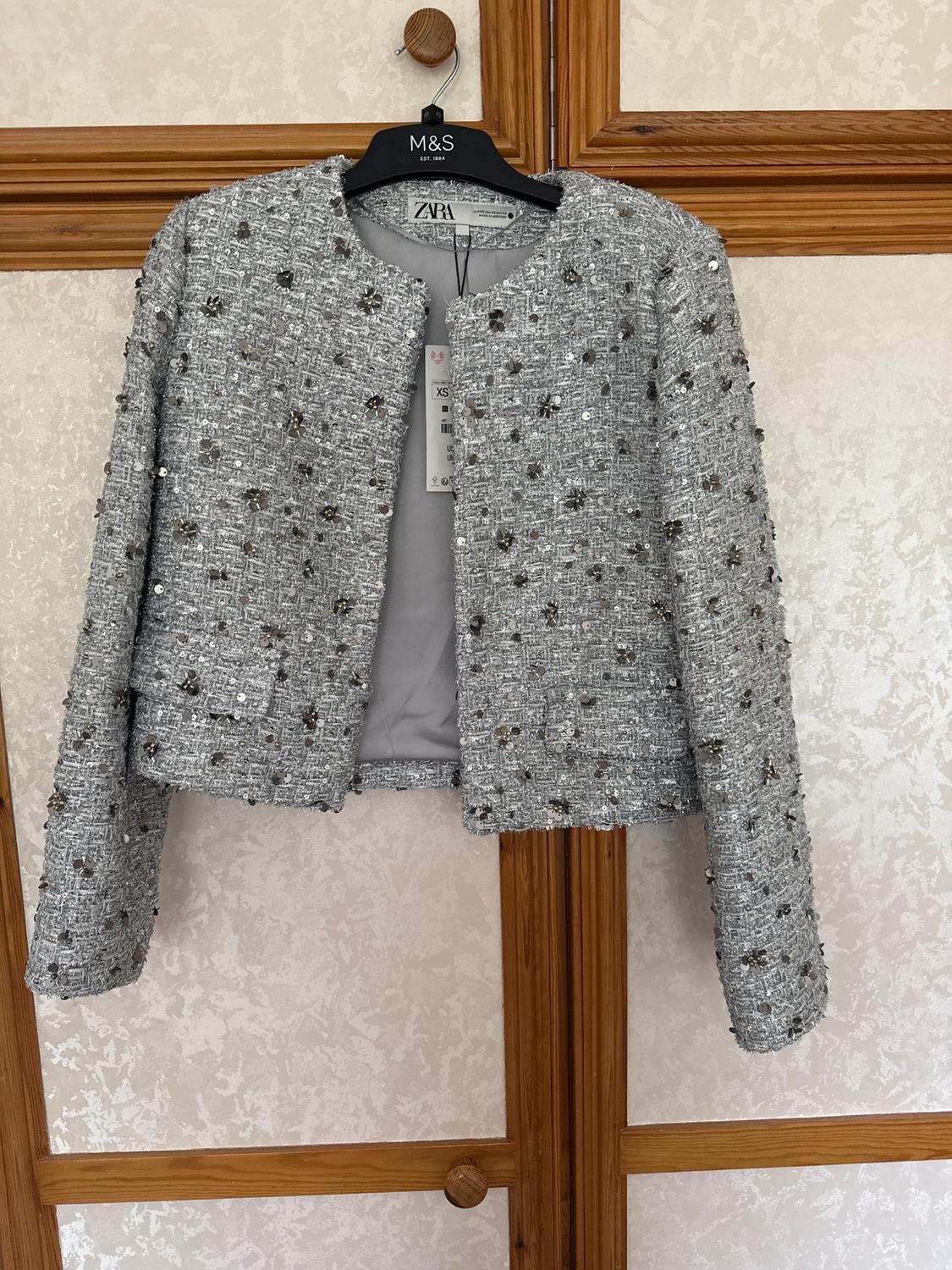 Textured grey sequin jacket hanging on an M&S hanger. Visible ZARA brand size XS label. Light wood background.