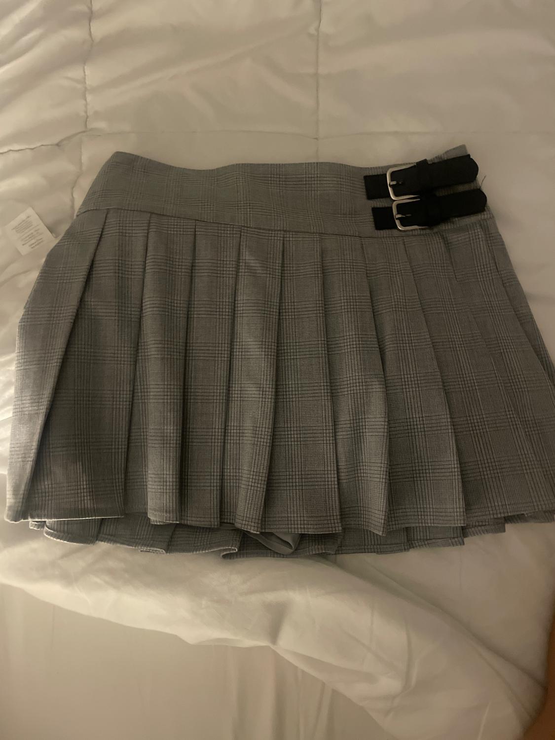 Grey checked mini skirt with pleats and two black buckles, on a white bedspread. Label visible on the left.