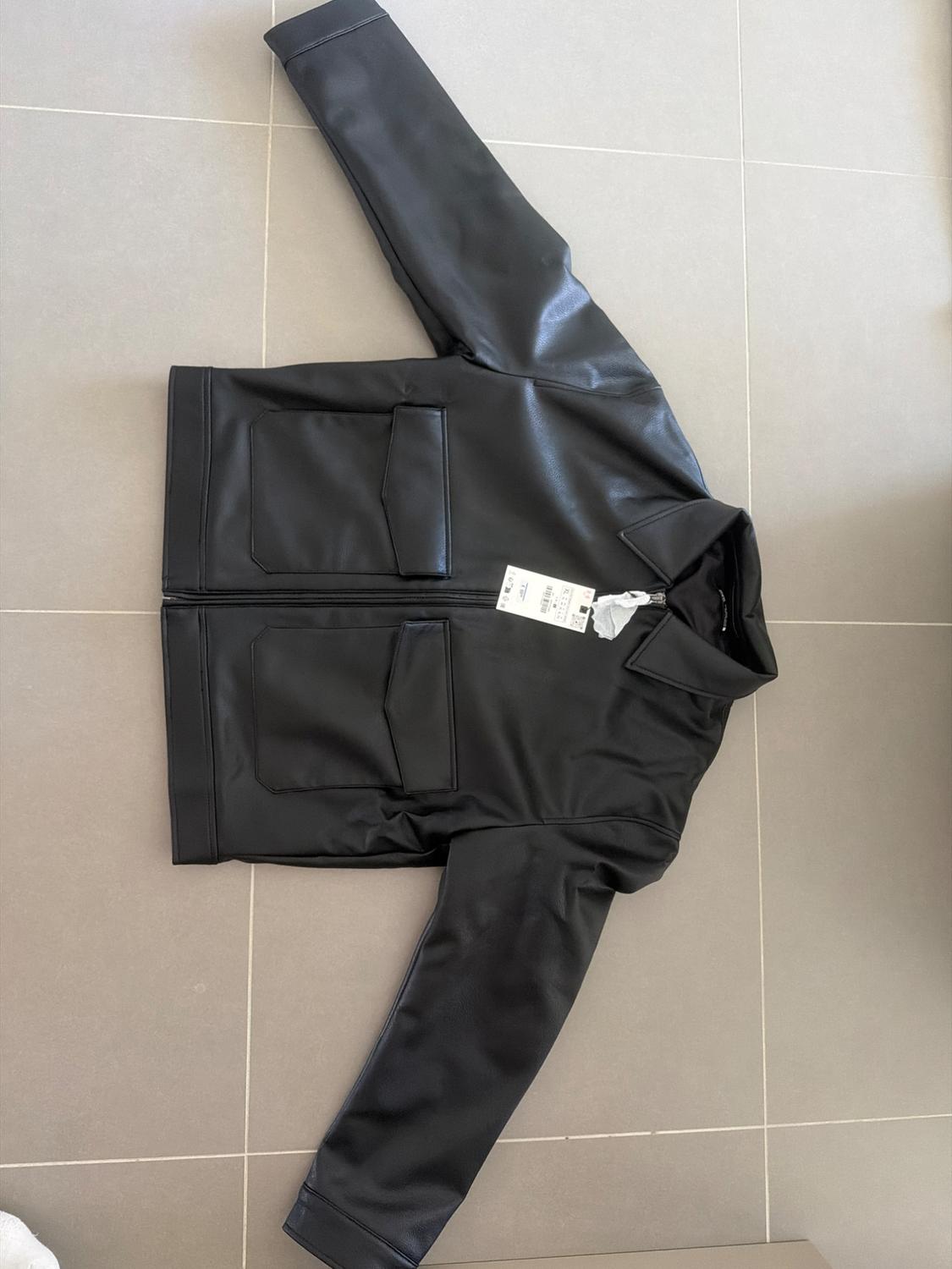 Black jacket with a collar, zip and front flap pockets, laid out on a light-tiled floor.