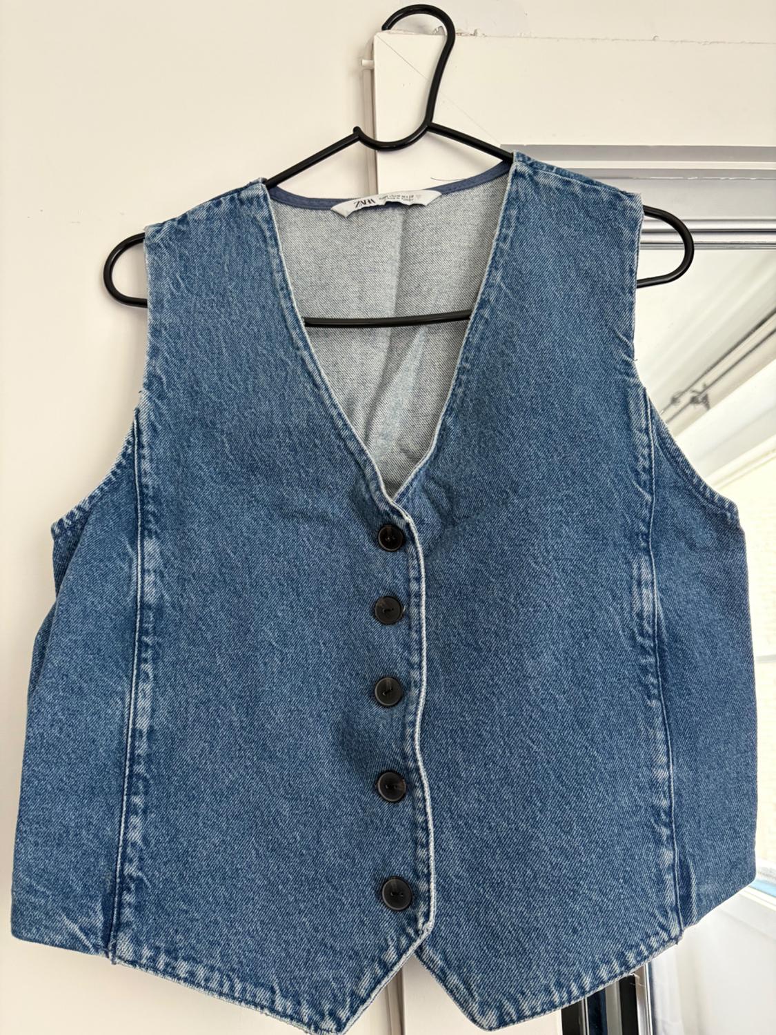 Blue denim waistcoat with a V-neck and buttons, hanging on a black hanger against a white door. Label with the text ZARA.