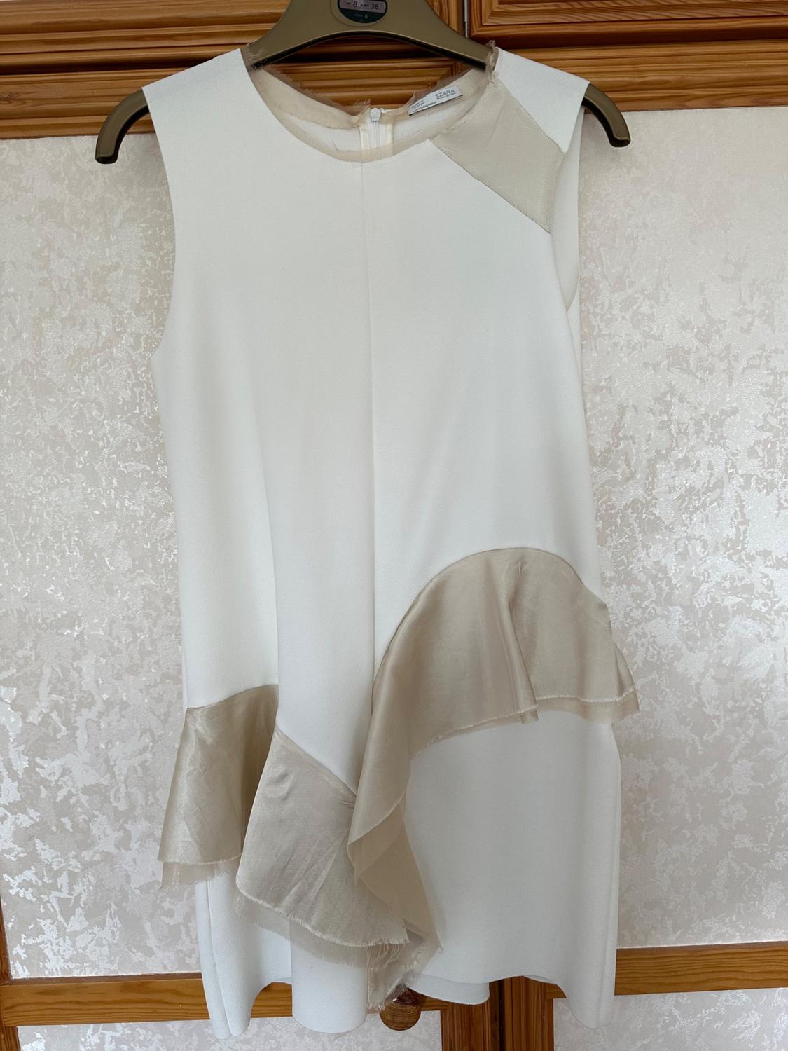 Front view of white sleeveless dress with beige frills on the hem and round neckline.