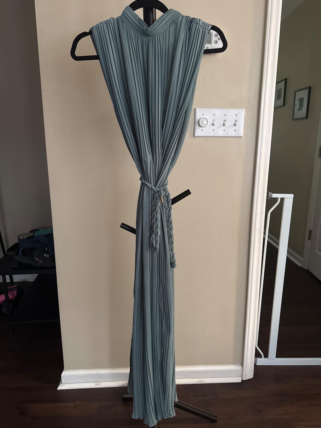 Blue pleated dress hanging on a black hanger in front of a beige wall, with a braided belt at the waist.