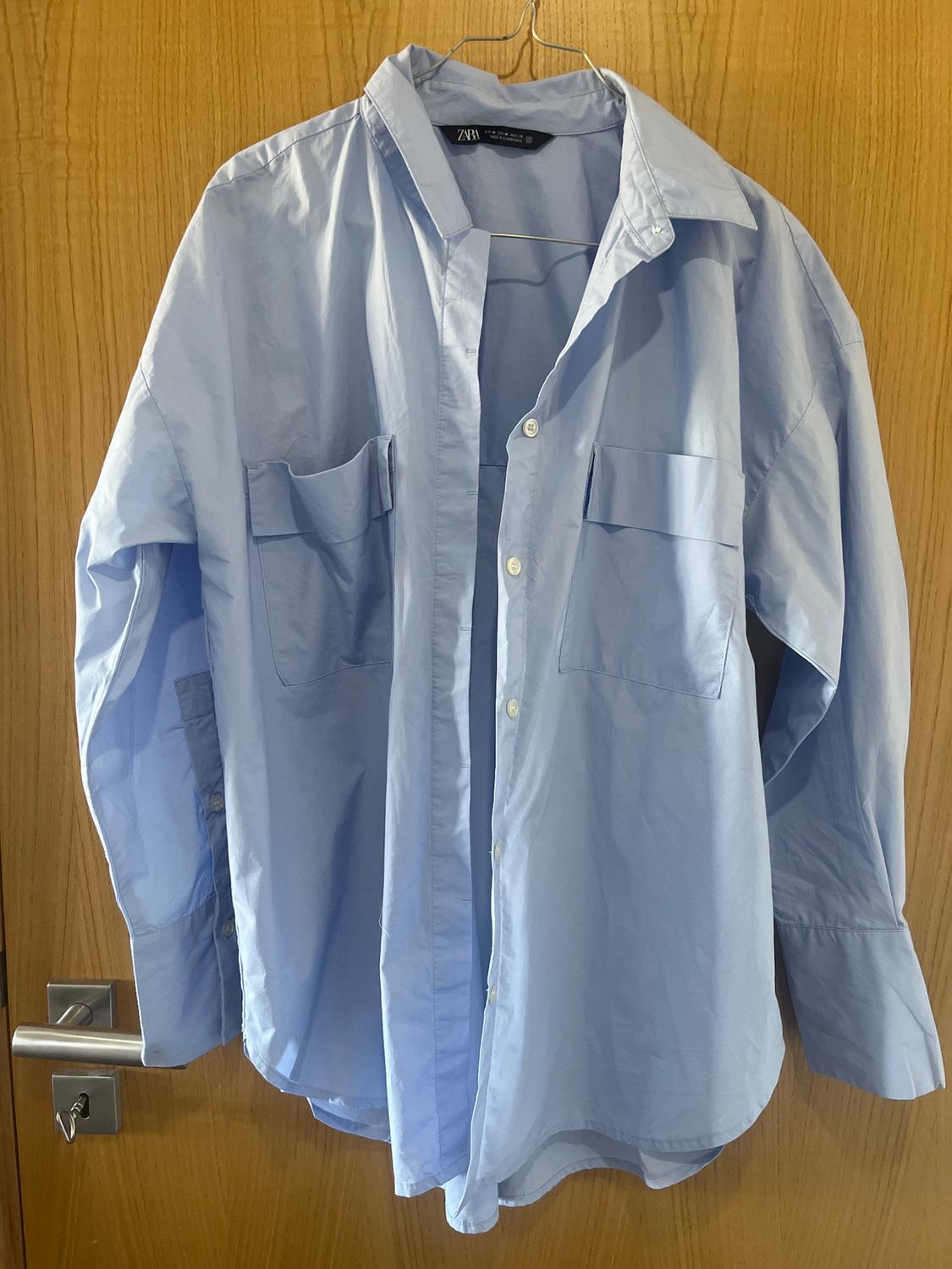 Light blue, long-sleeved shirt hanging on a hanger over a wooden door. The label reads "Zara".