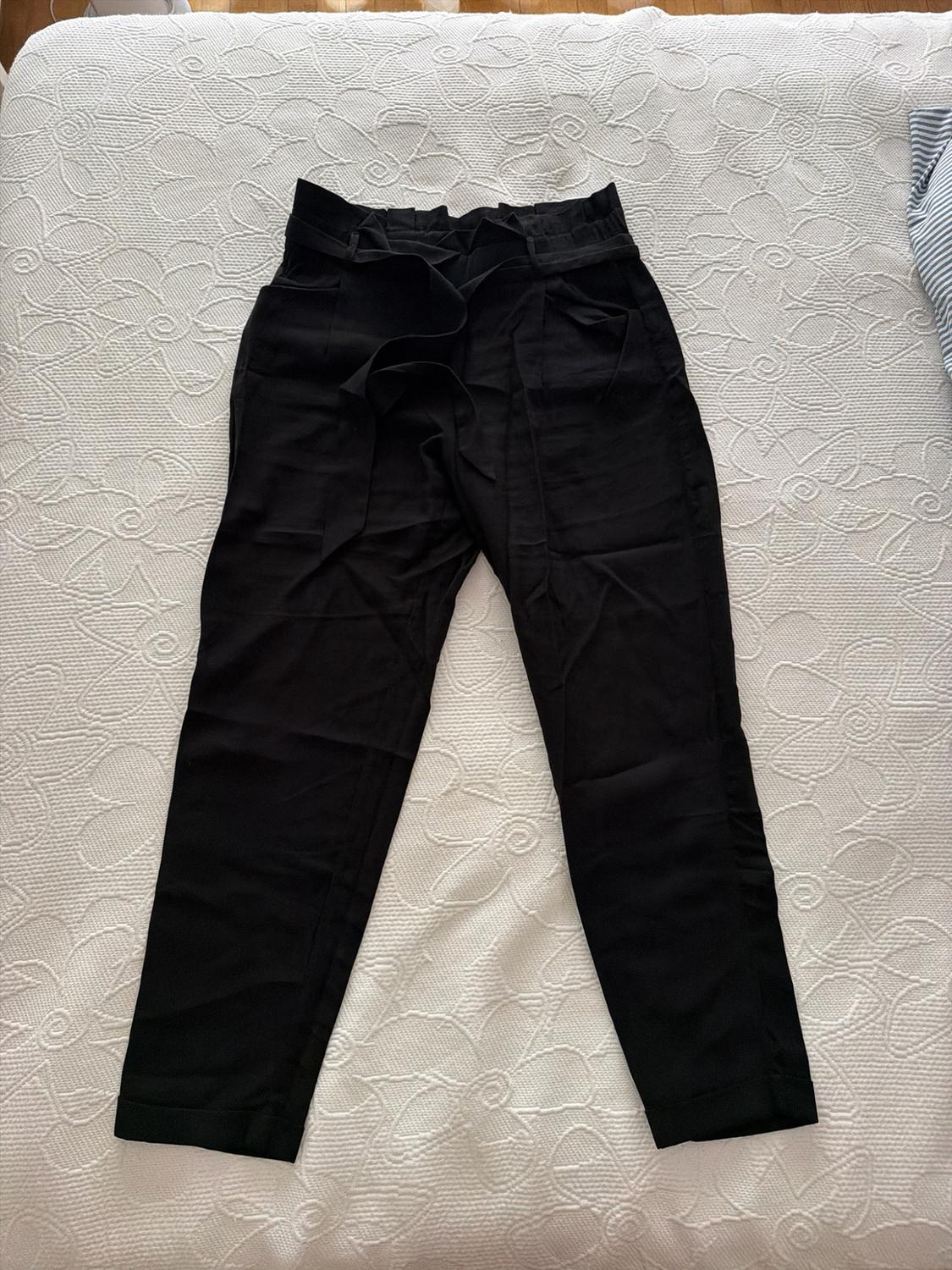 Black high-waisted gathered trousers with a tie, laid out on a white bedspread with a floral texture in a relaxed setting.