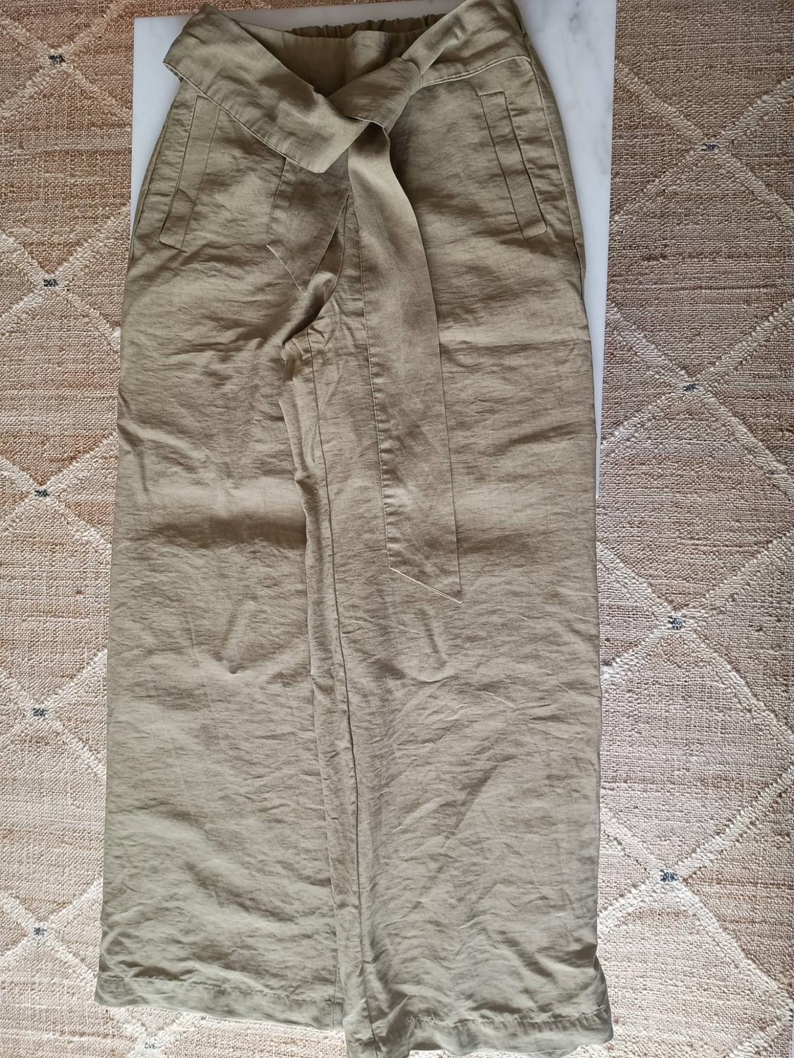 Wide-leg khaki trousers with a tie belt, arranged on a white surface and a textured diamond-patterned rug.