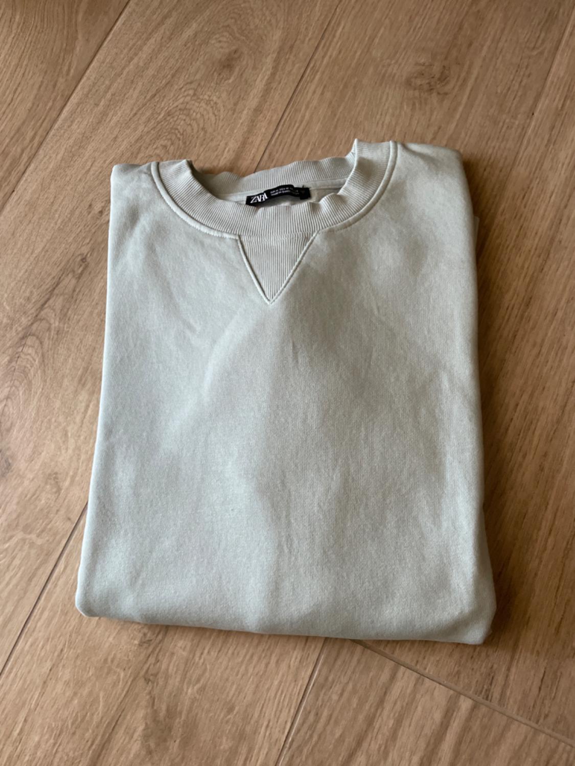 Light grey sweatshirt folded on a wooden floor. Round neck with ribbed V-detail. Label with the text ZARA. Relaxed composition.
