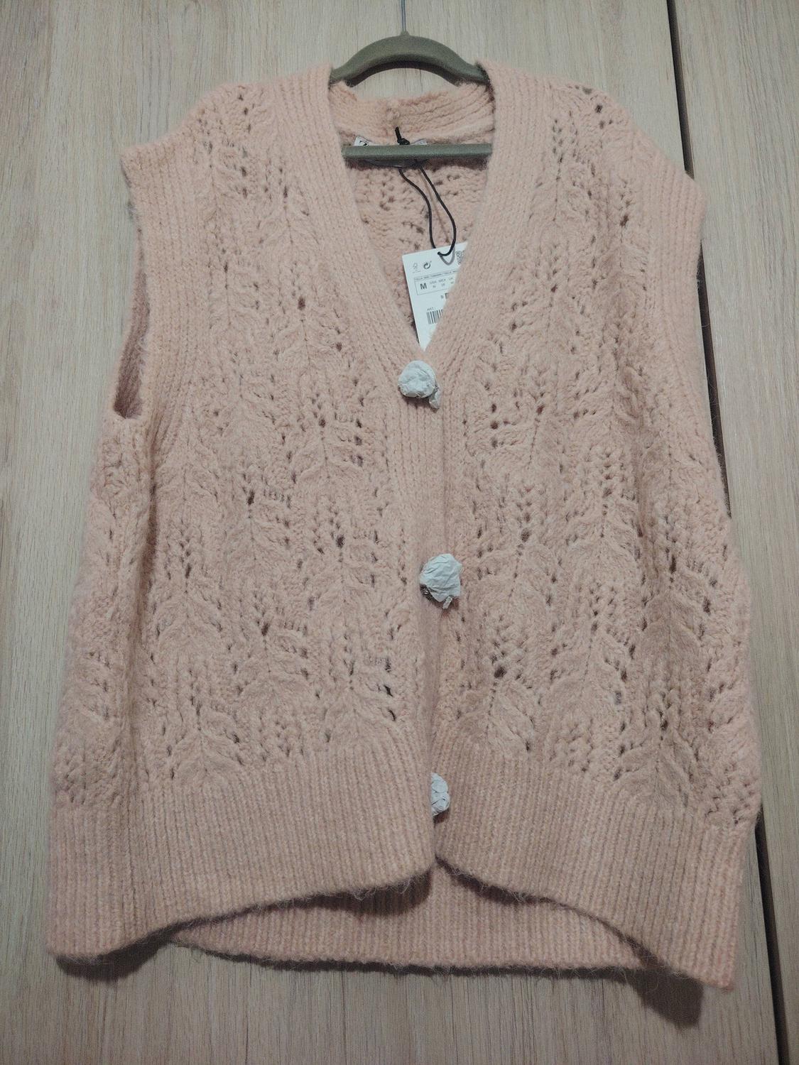 Light pink knitted waistcoat with white buttons, hanging on a hanger against a light wood background. Visible label with size M.