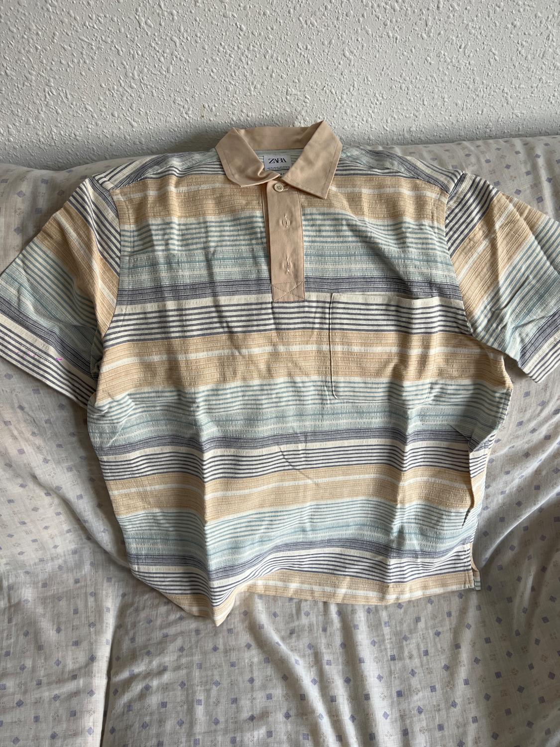 Horizontal striped shirt in shades of beige, blue, and grey on a bed with a white bedspread. Visible label on the collar.