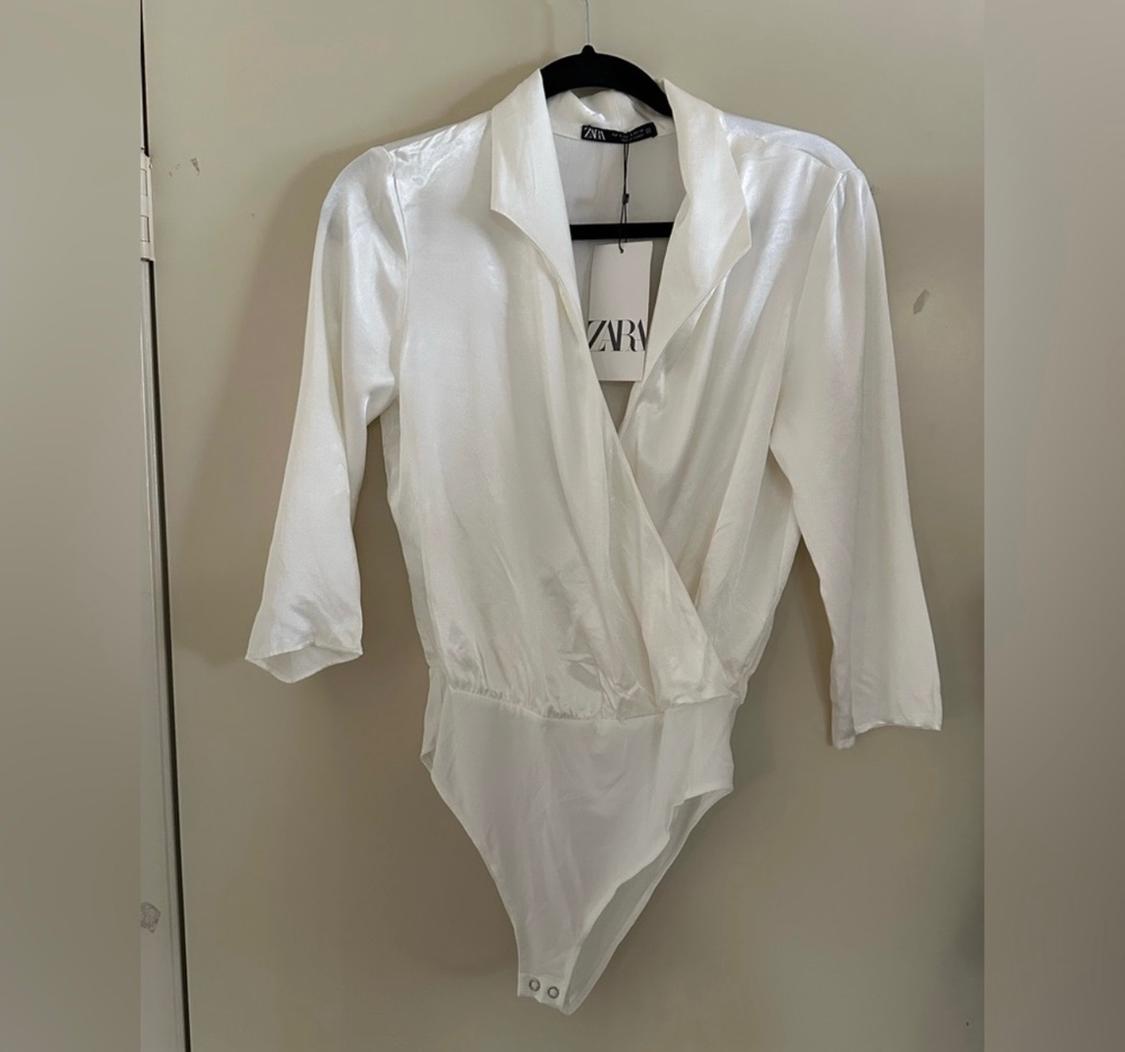 White long-sleeved bodysuit hanging on a black hanger. Visible label with the text "ZARA". Beige wall background.