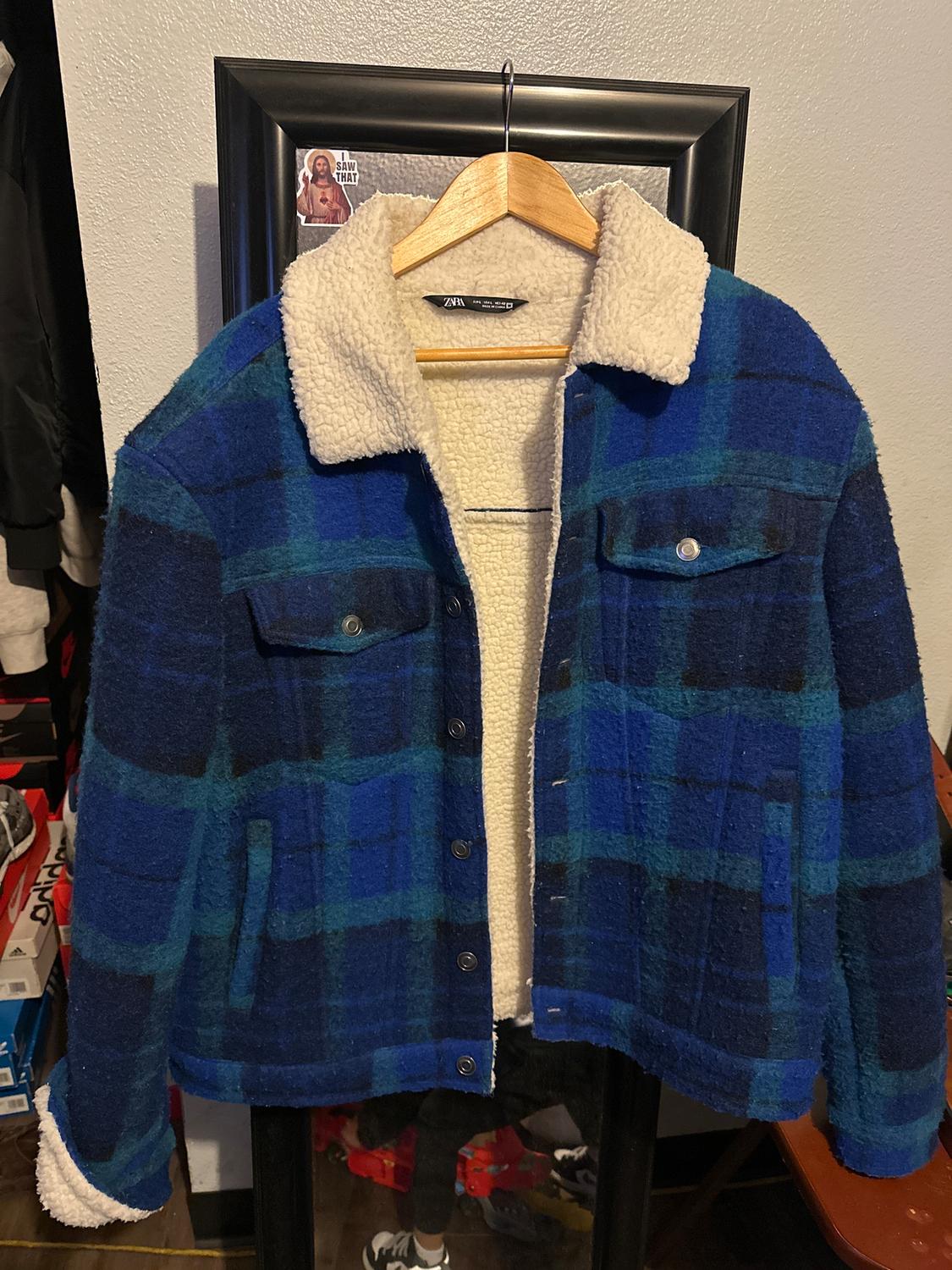 Blue and green checked jacket with white fleece lining, hanging on a wooden hanger in front of a grey wall.