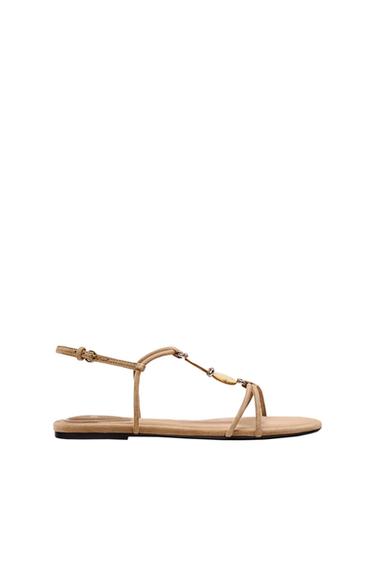 Beige flat sandal with thin straps, a circular metallic detail on the instep and an ankle buckle fastening, on a white background.