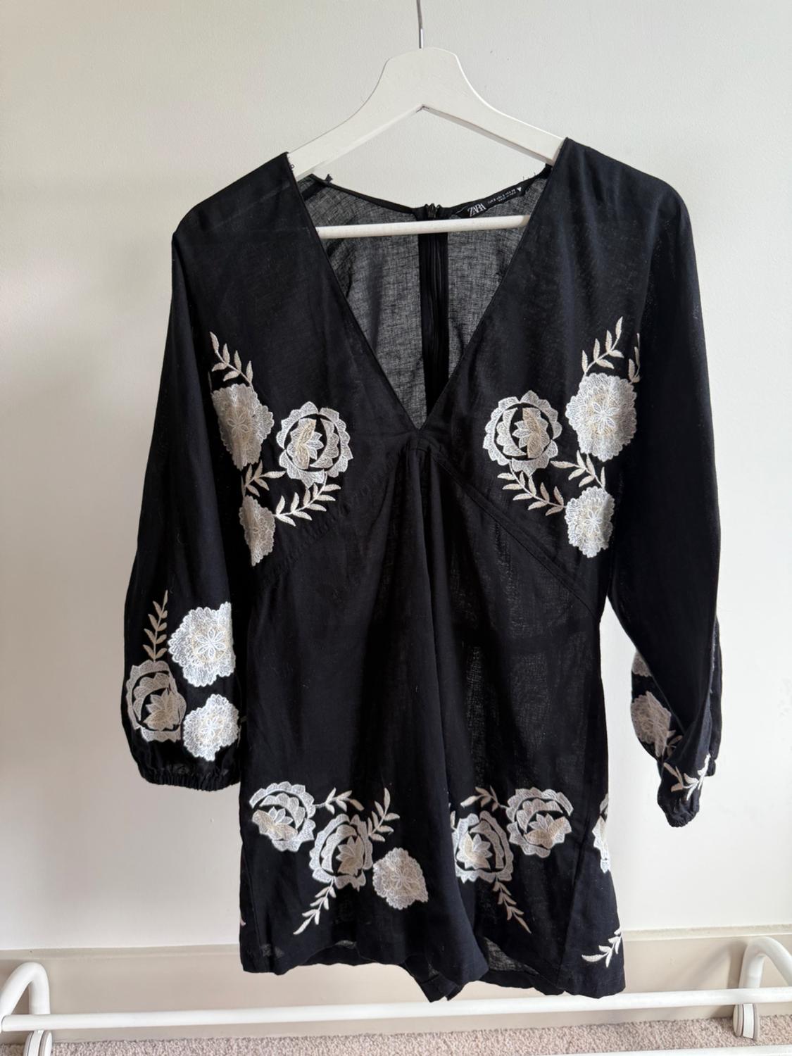 Black blouse with white floral embroidery on the sleeves and front, hanging on a white hanger against a light background.