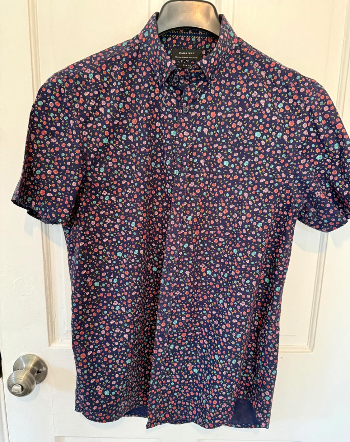 Short-sleeved shirt with a floral print in red and blue tones on a dark blue background, hanging on a white door.