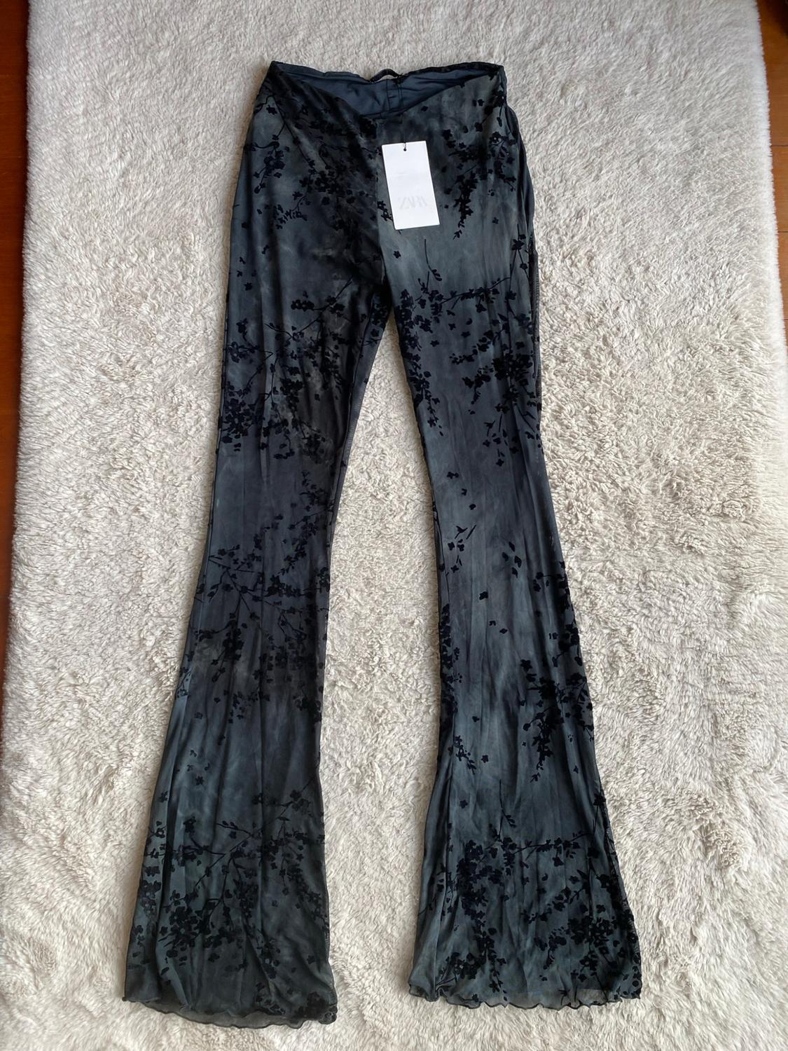 Dark grey flared trousers with a black floral print and ZARA label, laid out on a white rug with a soft texture.