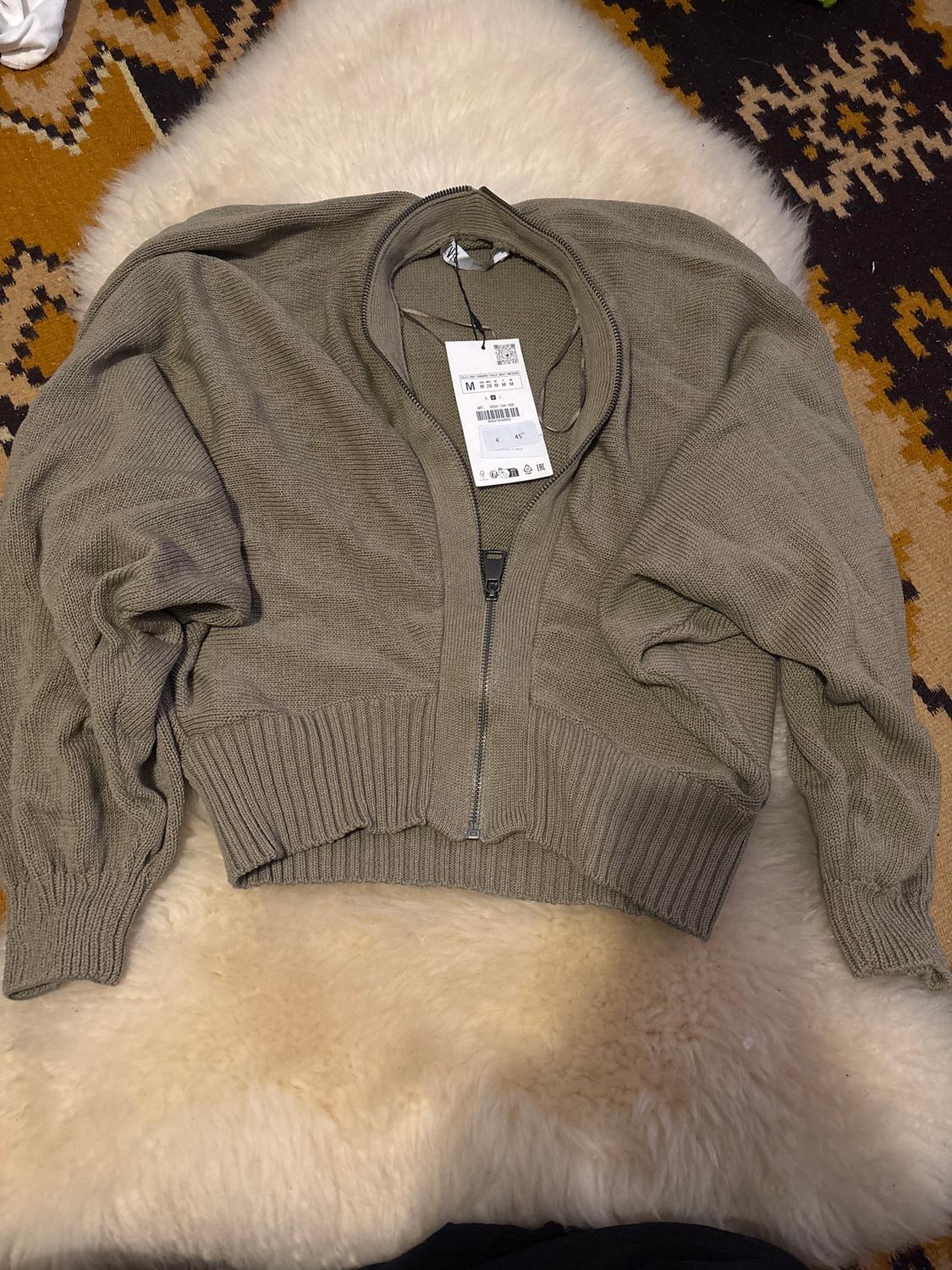 Grey zip-up jacket with size M label on a white and brown geometric patterned rug.