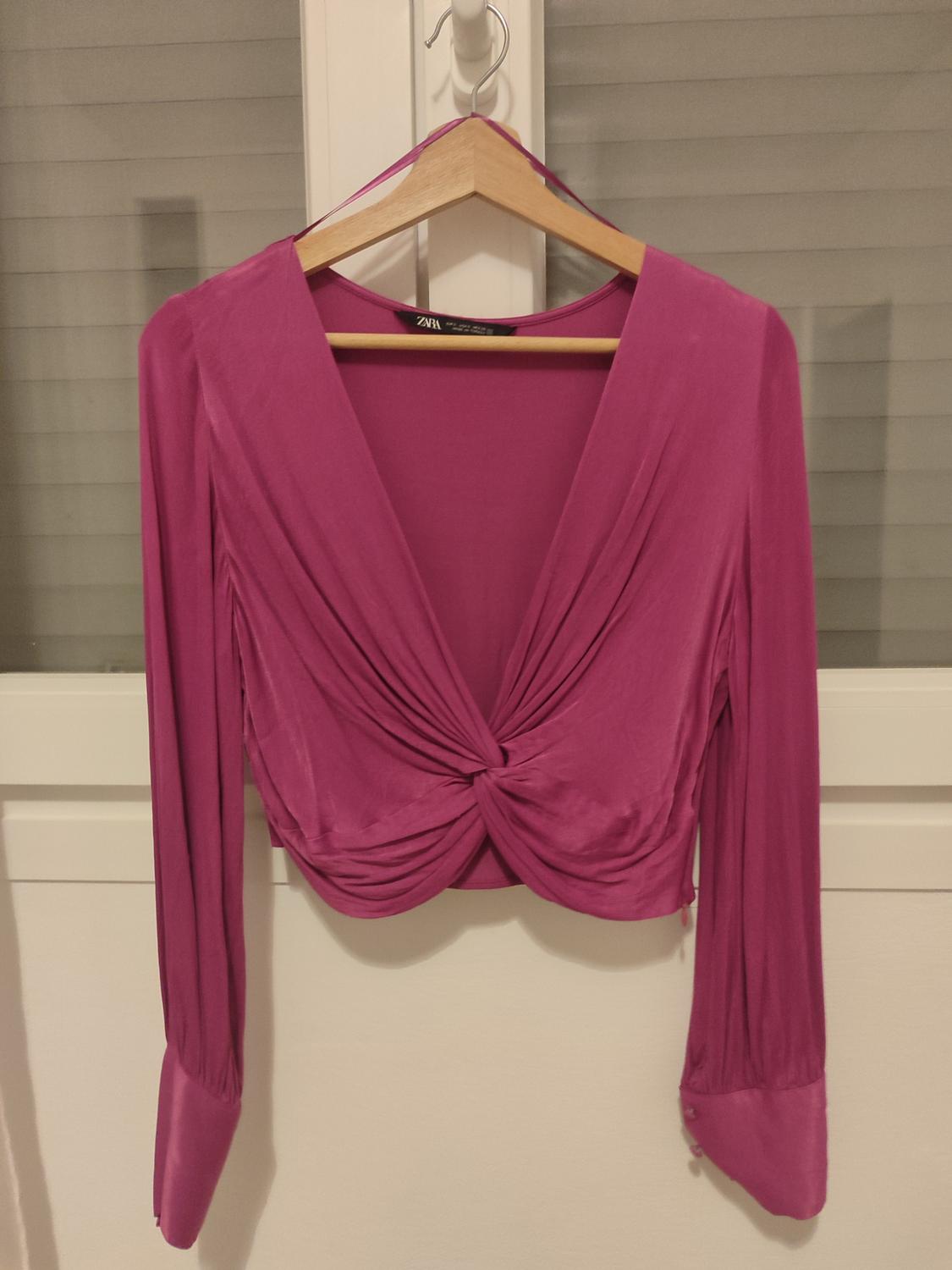 Fuchsia long-sleeved blouse with a V-neck and front knot, hanging on a wooden hanger in front of a white window.