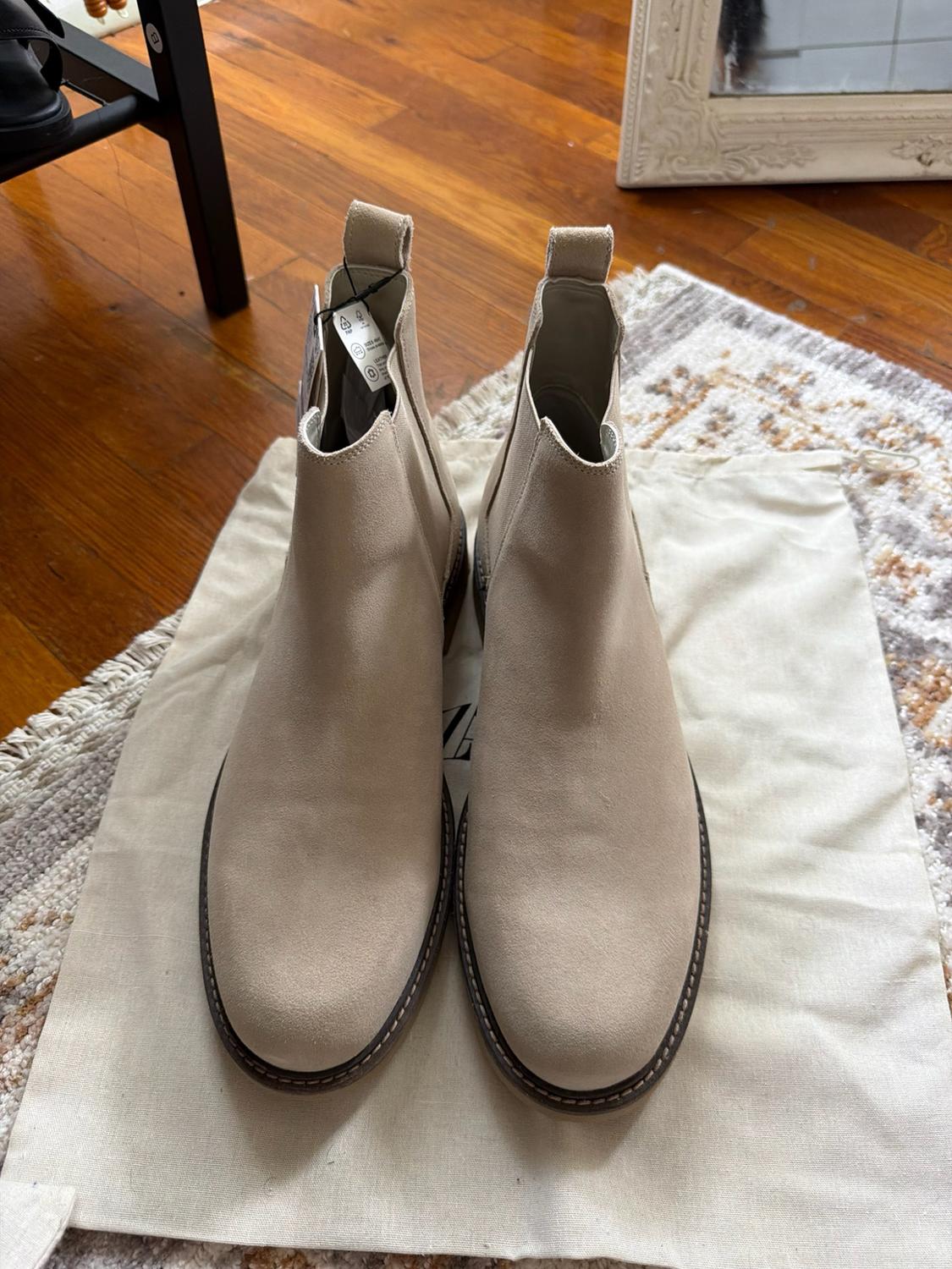 Beige suede Chelsea boots on a white fabric bag with the text ZA,
resting on a rug and a wooden floor.