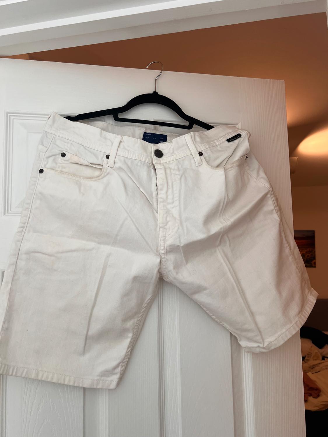 White denim shorts hanging on a black hanger over a white door in a room. Visible text: ZARA MAN.