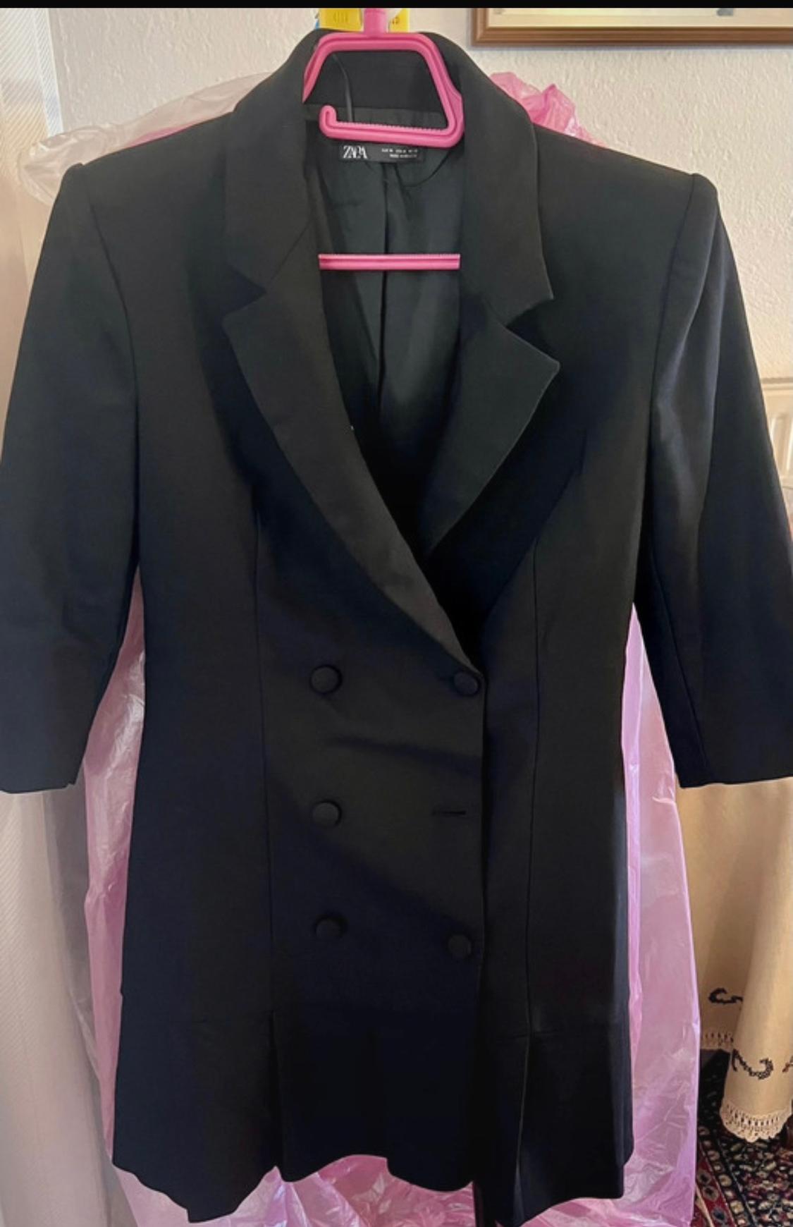 Black double-breasted blazer dress with buttons and pleated hem on a pink hanger. ZARA label. Casual indoor setting with plastic cover.