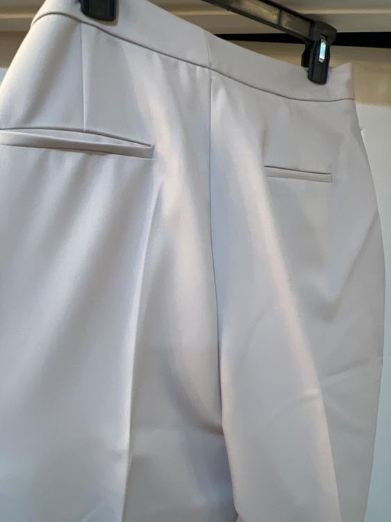 White pants hanging on a hanger, showing details of back pockets and pleats in a well-lit indoor setting.