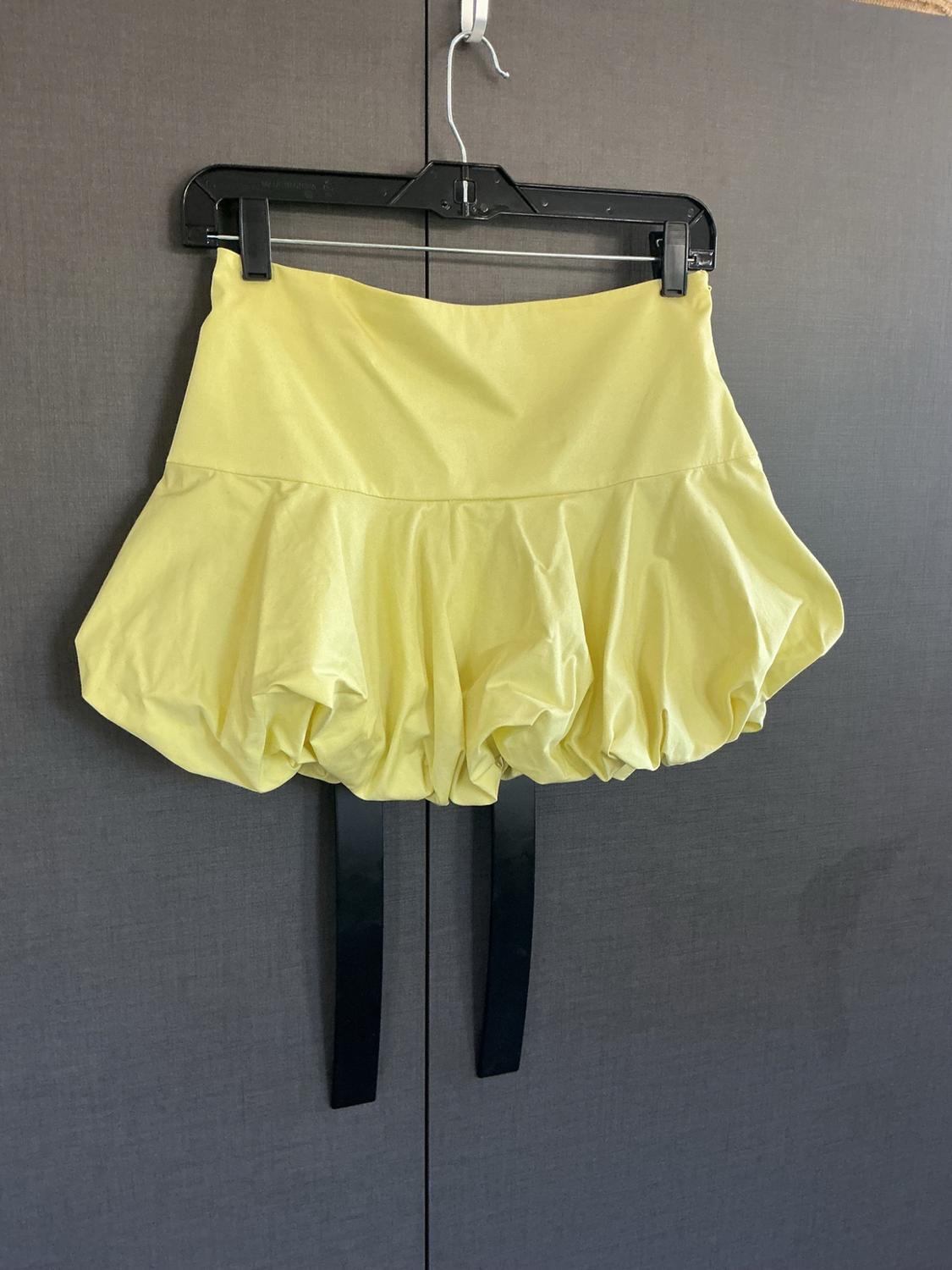 Short yellow balloon-design skirt hanging on a black hanger in front of a dark closet, with two black ribbons hanging from the hem.