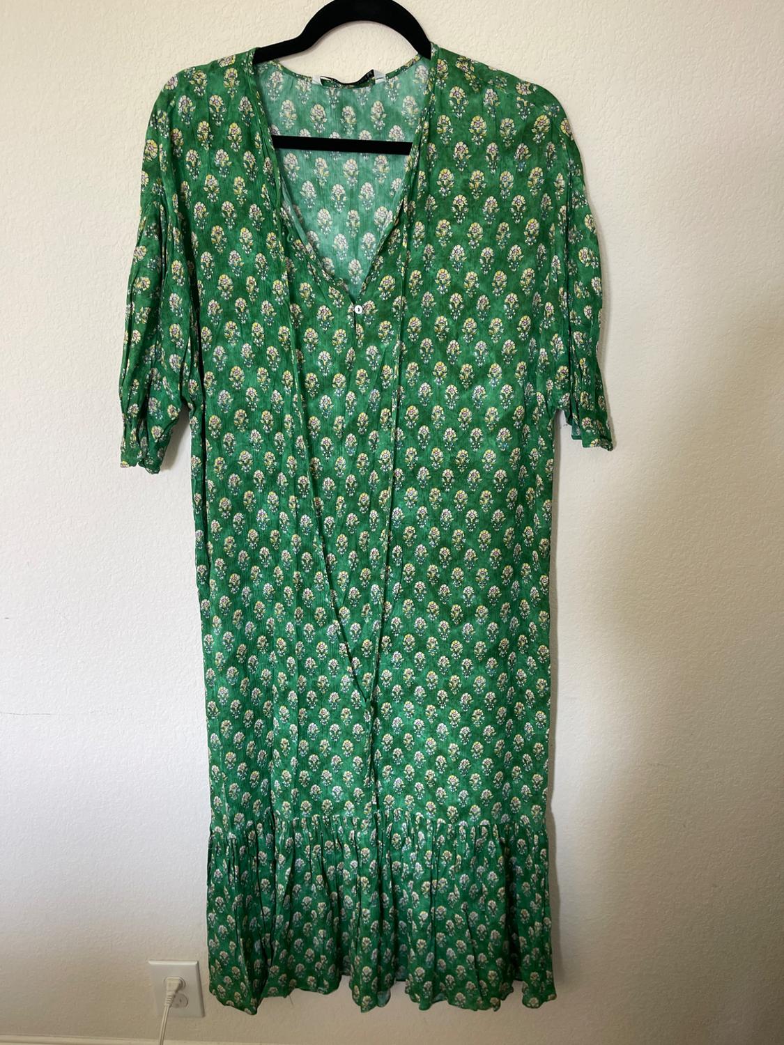 Green dress with white and yellow floral print, short sleeves and V-neck, hanging on a black hanger against a white wall.