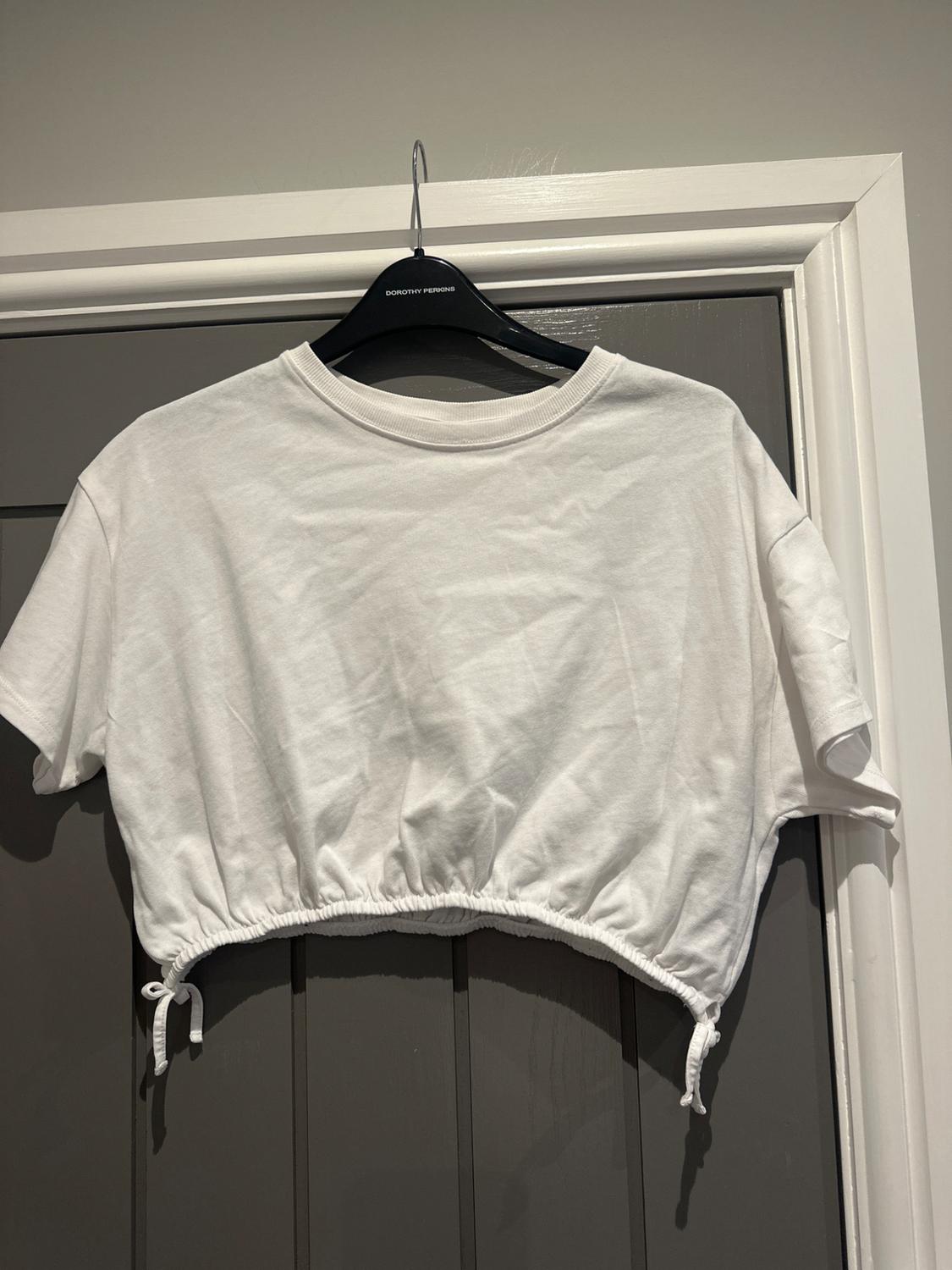 White cropped T-shirt with drawstring hem, hanging on a black hanger that says "Dorothy Perkins" over a grey door.