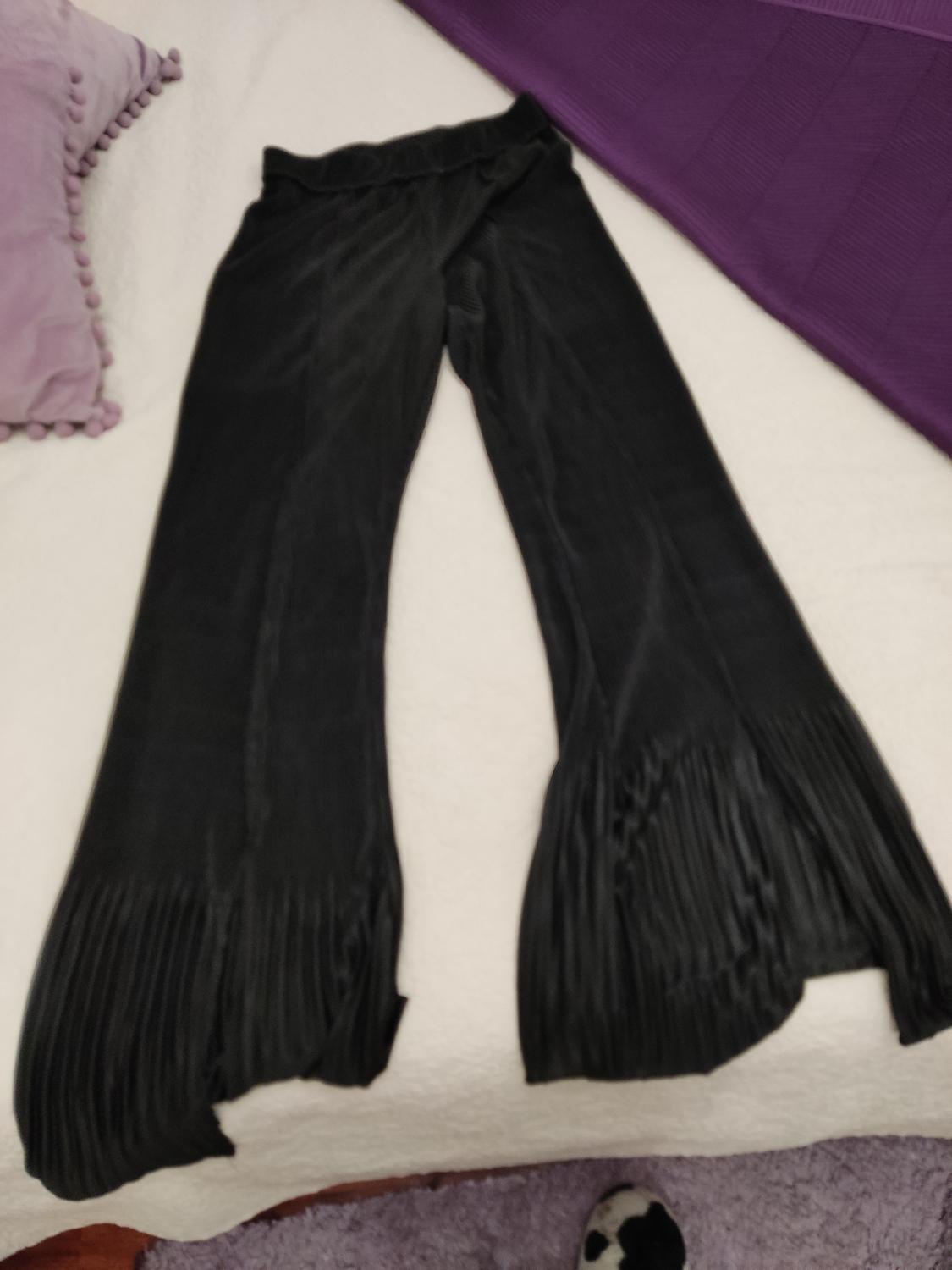 Pleated black trousers on a white bed with purple cushion and blanket. Part of a rug and slipper visible.