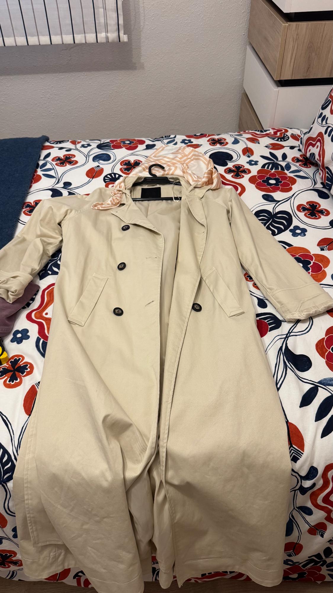 Beige trench coat with black buttons on a bed with a red and blue floral bedspread. Striped white curtain in the background.