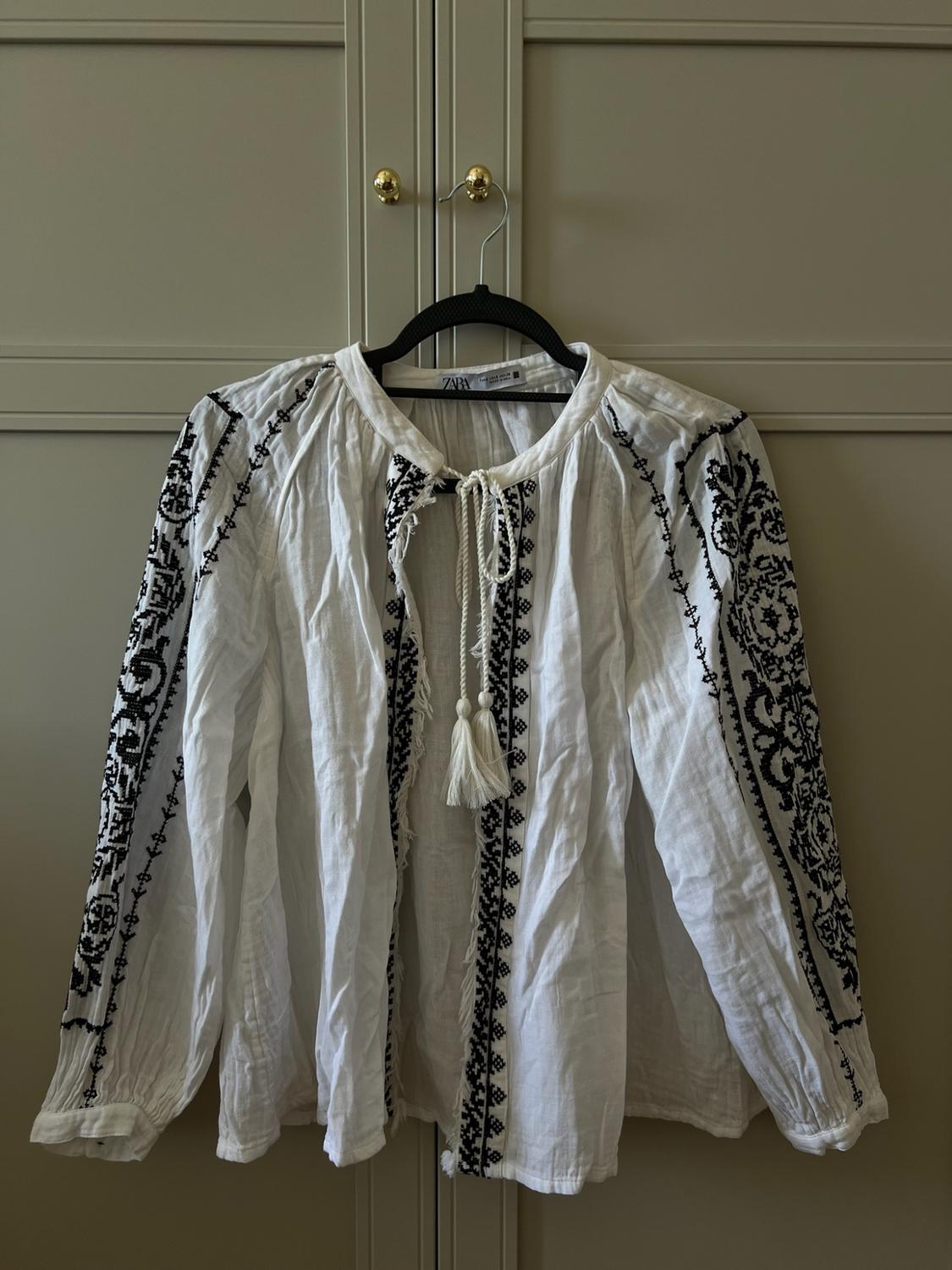 White blouse with black embroidery on the sleeves and front, ties with tassels. Hanging on a hanger in front of a wardrobe. Text: ZARA.
