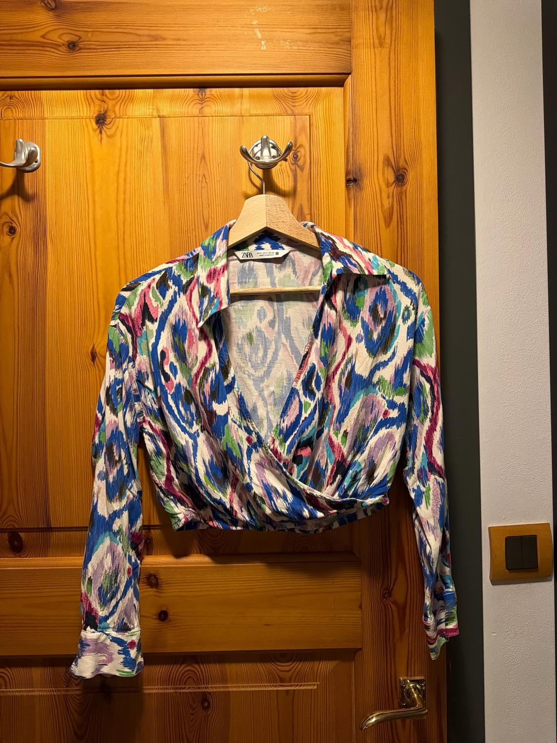 Cropped long-sleeved wrap blouse with an abstract blue, green and pink print, hanging on a wooden door. ZARA label.