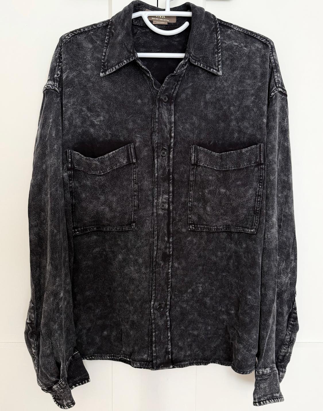 Black distressed denim shirt with two front pockets and buttons, hanging on a white hanger against a light background.