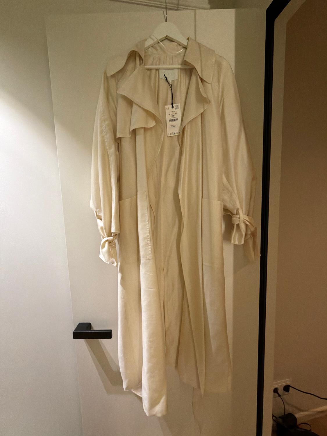 Beige coat hanging on a white hanger, with a visible tag, on an open door. Bright and minimalist setting.