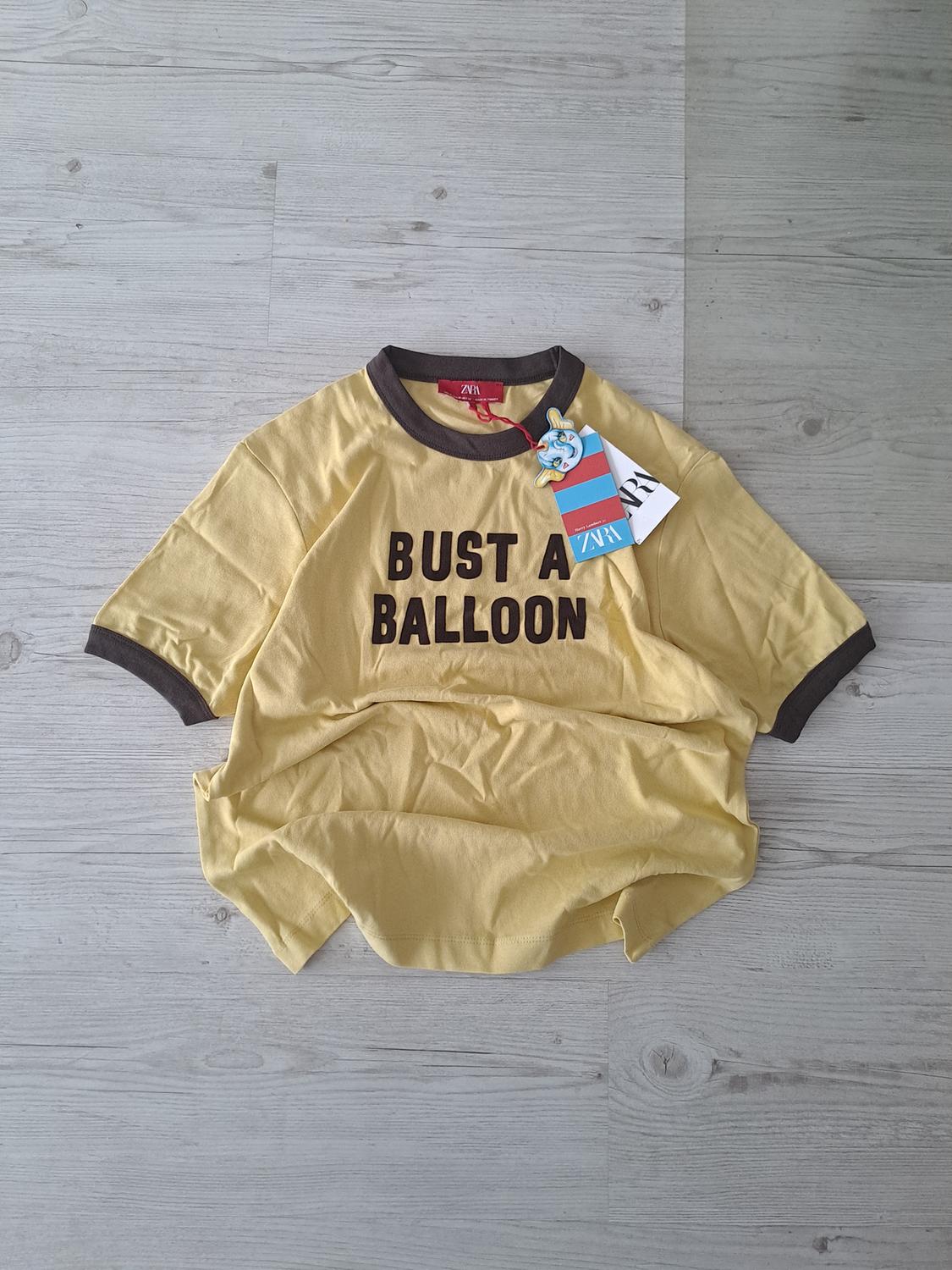 Yellow t-shirt with brown trim on a light wooden floor. Text "BUST A BALLOON" on the front. Brand label visible.