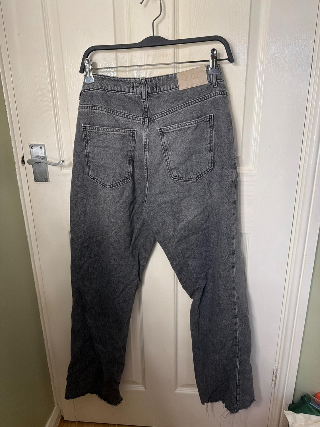 Grey denim jeans hanging on a black hanger over a white door. Back pockets and brown label visible.