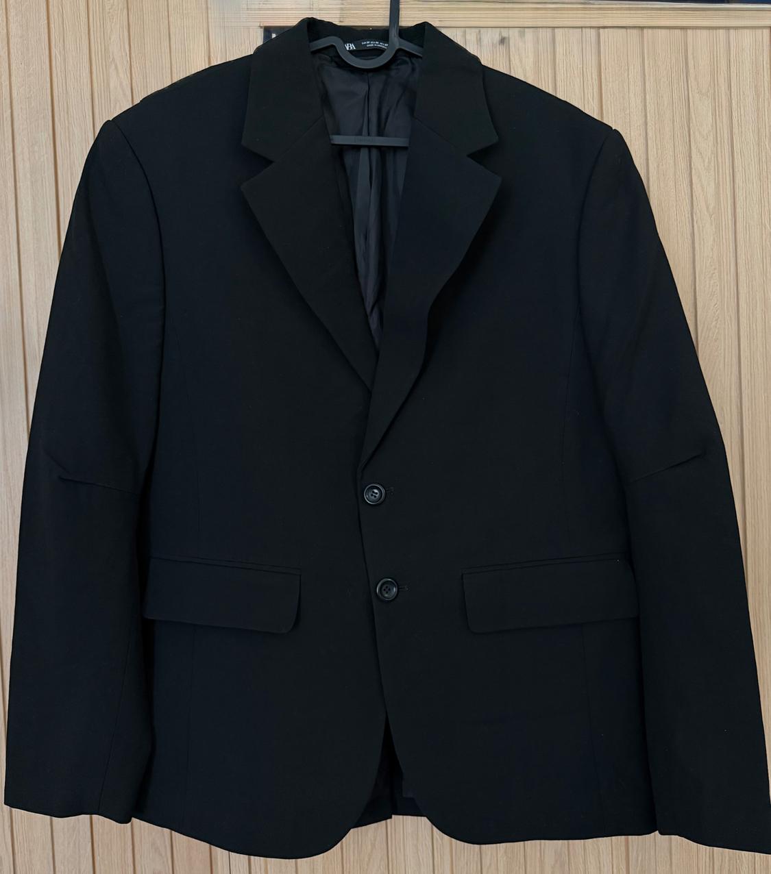 Black two-button blazer with flap pockets, hanging on a hanger against a light wood panelled background.