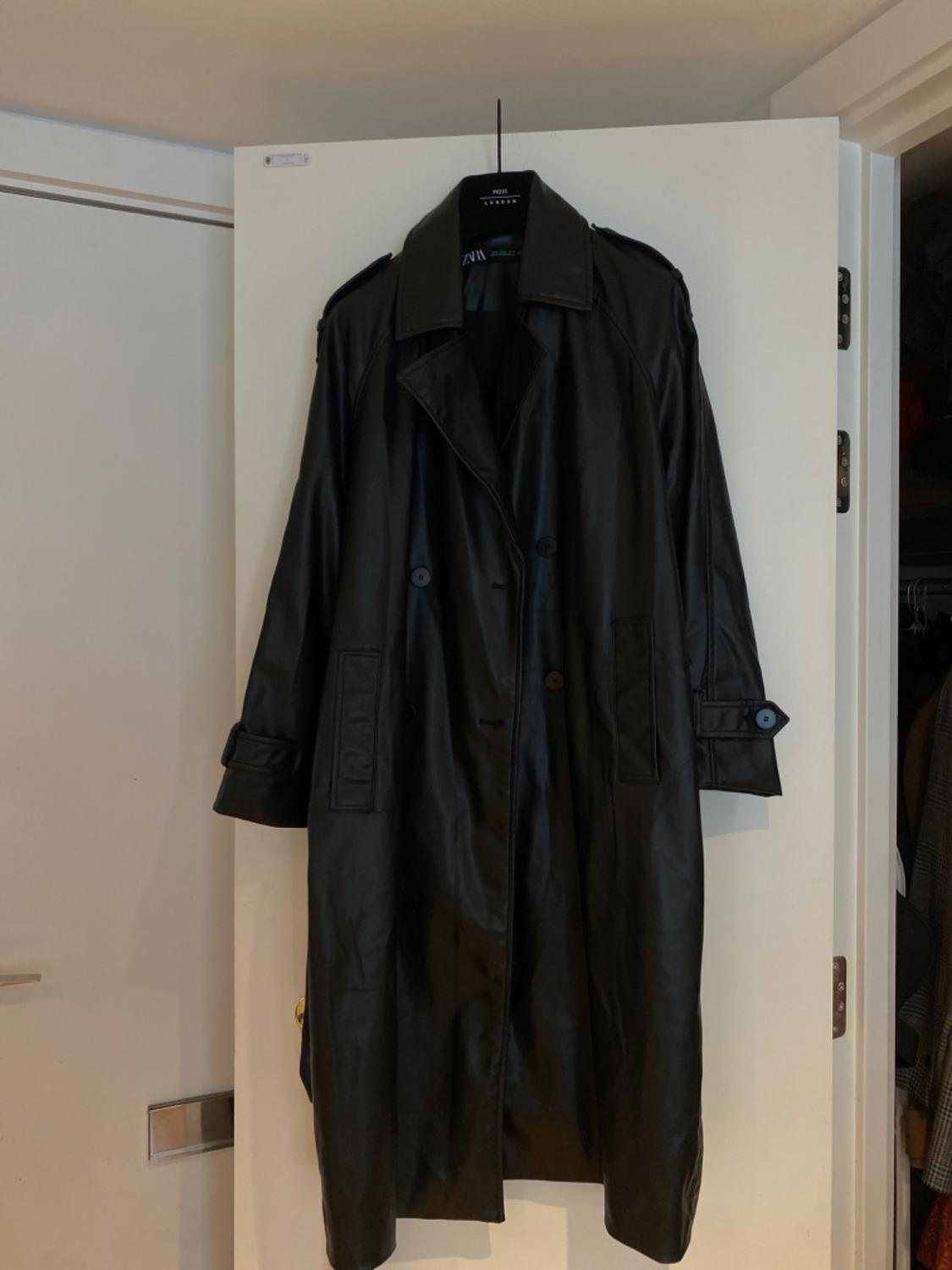 Long black coat hanging on a white door. It features a double-breasted design with buttons, wide lapels, pockets and cuff tabs.